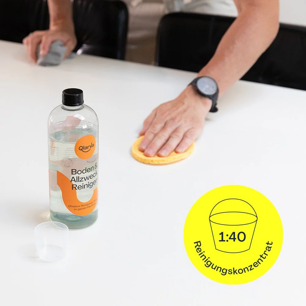 Person, die eine Tablette mit einem Reiniger und einem Schwamm auf einem weißen Tisch abwischt, mit einem Flasche Reinigungsmittel in der Nähe und einer gelben Beschriftung mit 1:40 Reinigerkonzentrat.
