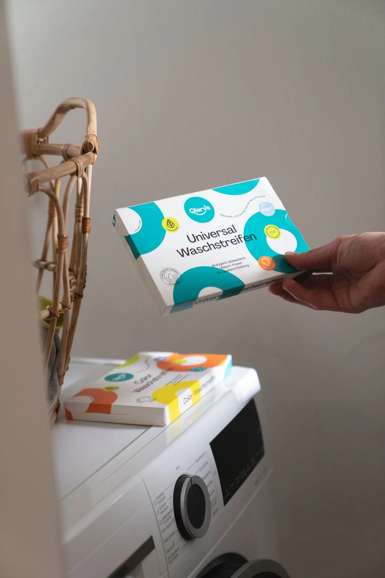 Eine Hand hält eine Packung mit Reinigungswaschstraßen auf einer Wäschemaschine, im Hintergrund ist eine zweite Packung auf einem Tisch zu sehen.