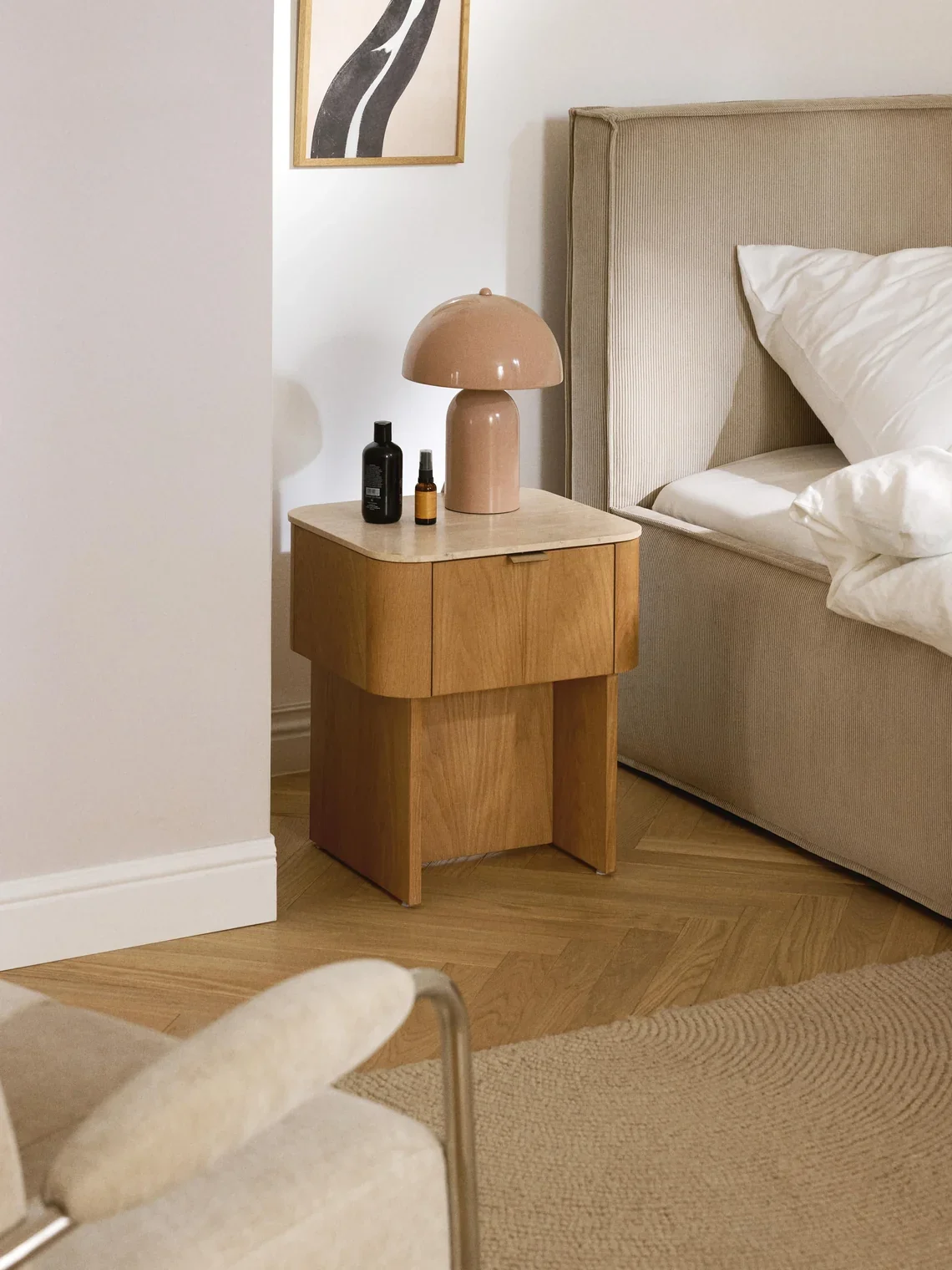 Nightstand with a beige mushroom-shaped table lamp, black and amber bottles, next to a beige upholstered bed with white bedding in a bedroom.