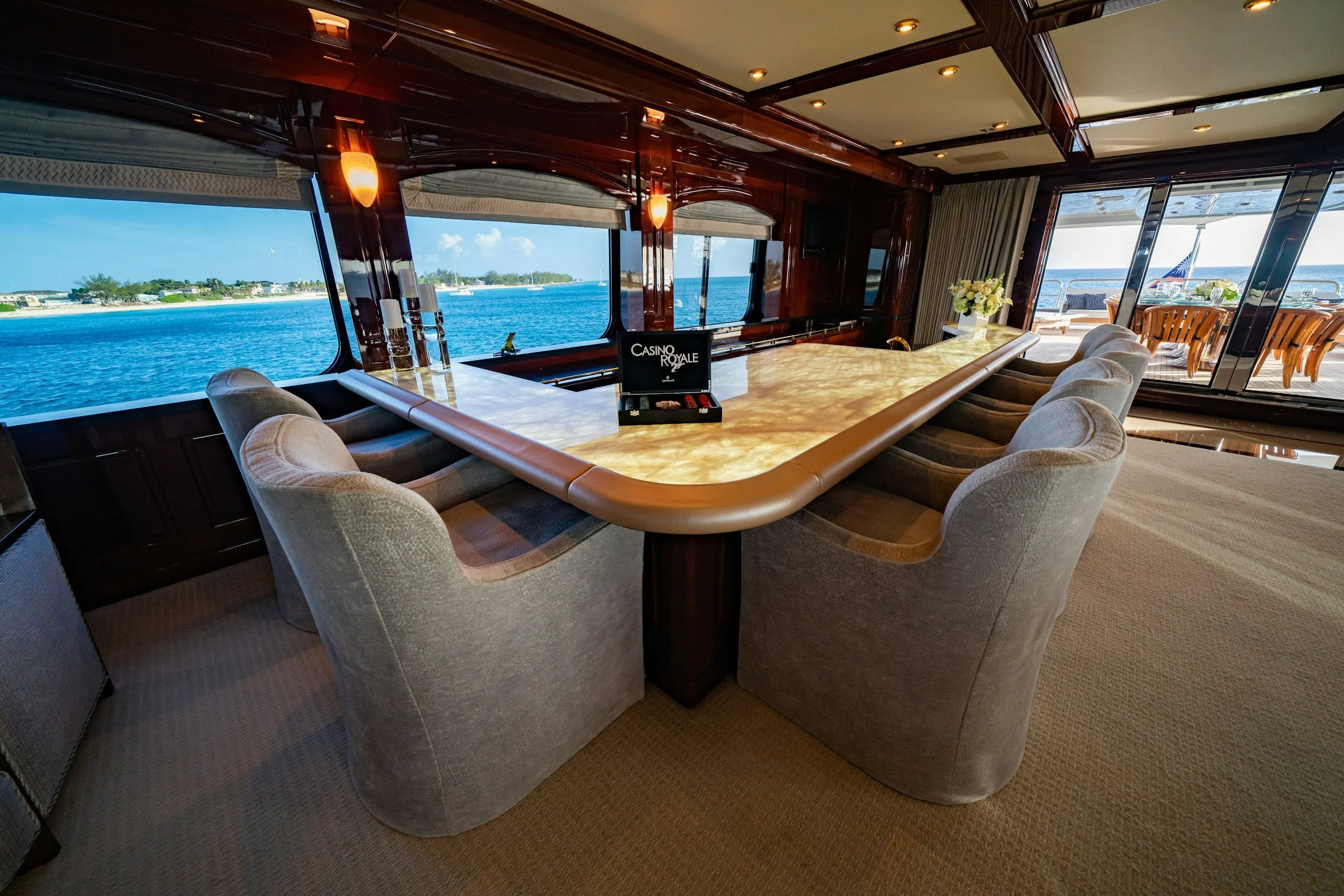 Inside of a luxurious yacht with a large marble table surrounded by beige upholstered chairs, overlooking the water through large windows, with an outdoor dining area visible through sliding glass doors.