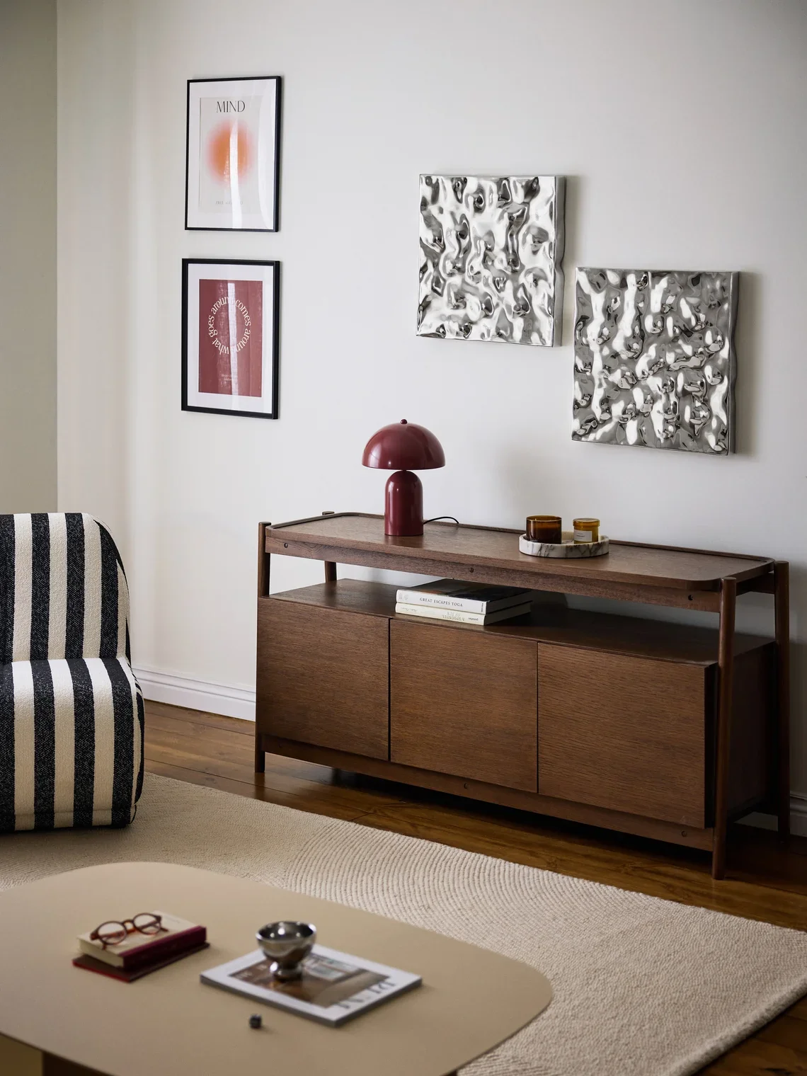 Modern living room corner with a wooden sideboard, a red table lamp, wall art, and a striped armchair.