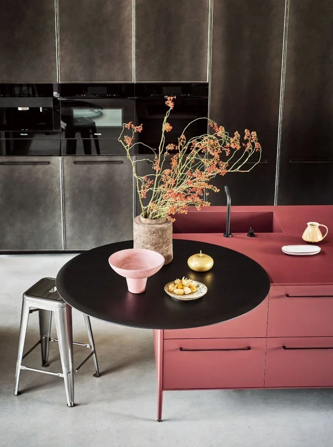 Modern kitchen with matte black and pink cabinetry, a black round table with decorative vases and a pink bowl, a metal stool, and a large potted plant with dried flowers.