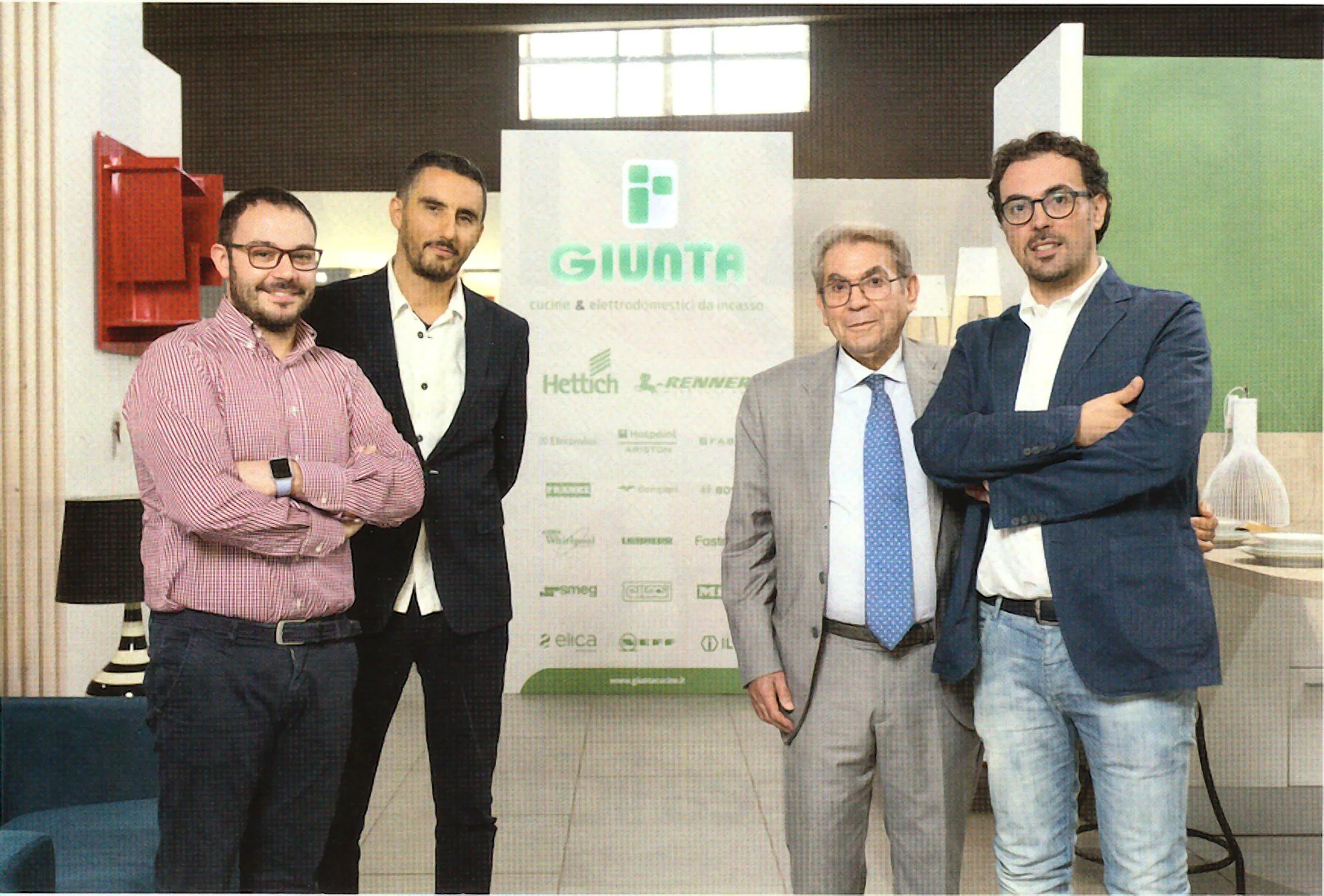 Five men standing in front of a sign with the word 'GIUNTA' and other text and logos, posing for a photo indoors.