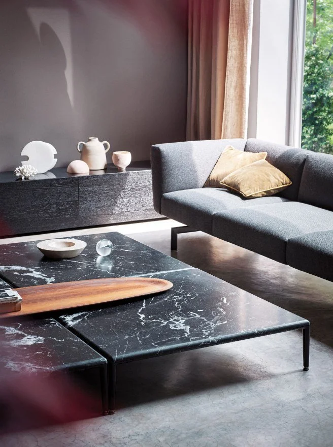 Modern living room with a gray sofa, gold pillow, black marble coffee table, and a black sideboard decorated with white and beige ceramics near a window with curtains.