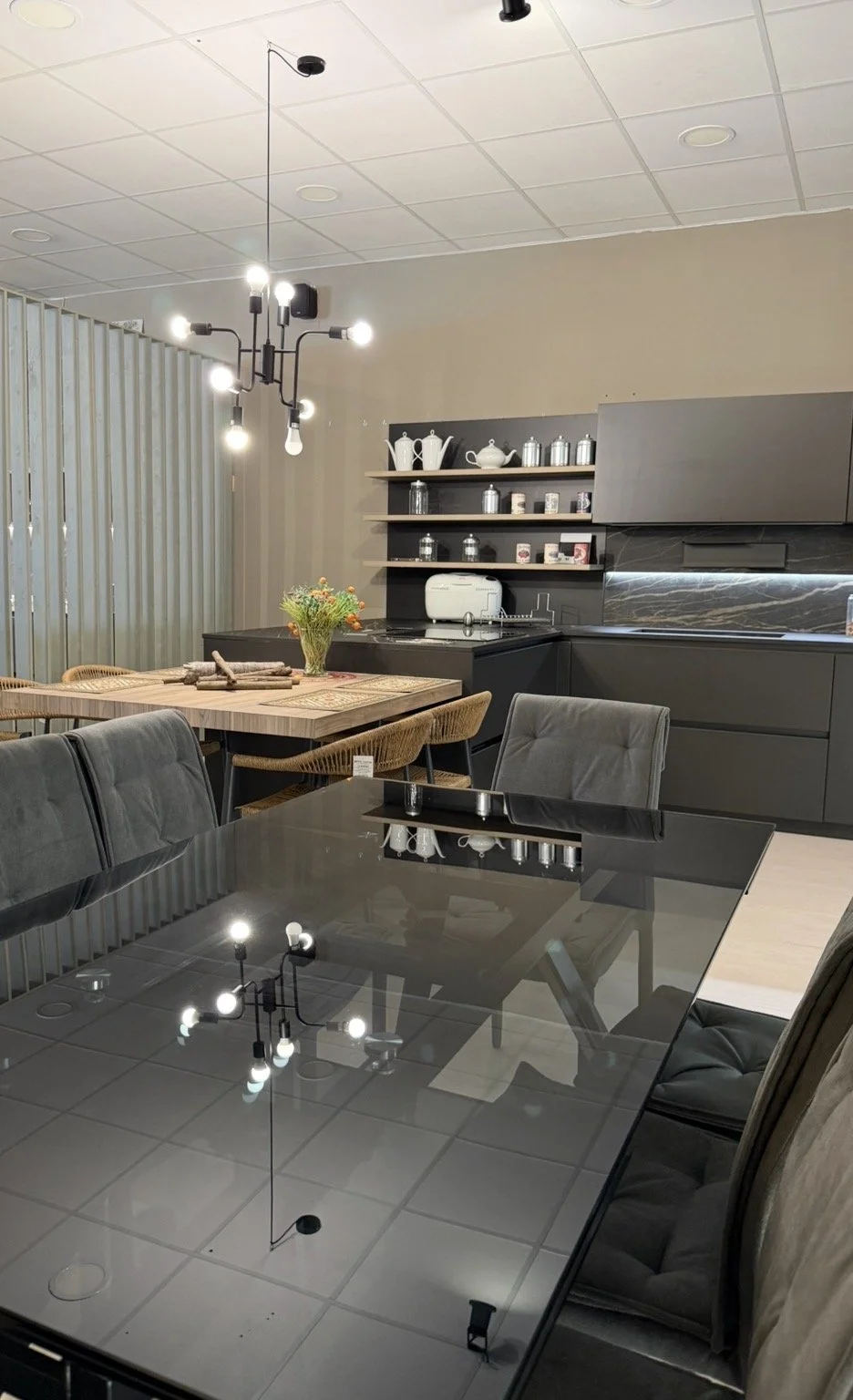Modern kitchen and dining area with black glass table, gray chairs, wooden dining table with flowers, and black and gray cabinetry with open shelves.