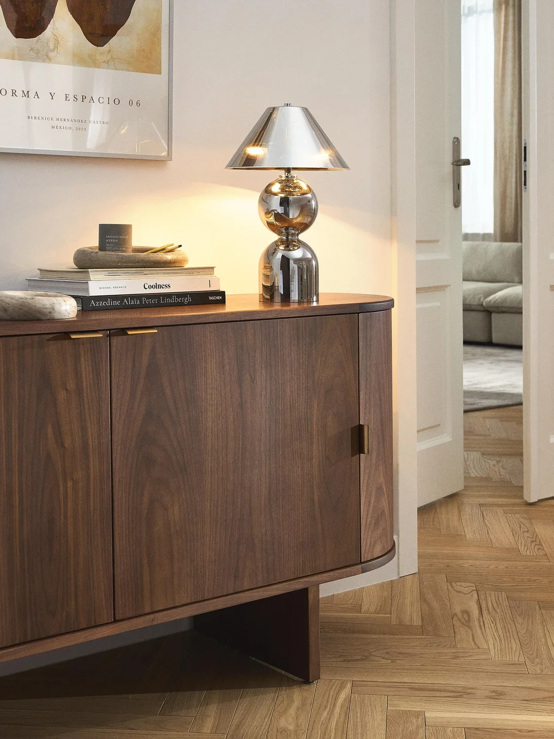 A wooden sideboard with a metallic lamp, books, and decorative items on top, in a living room with a partially open door leading to another room with a sofa.