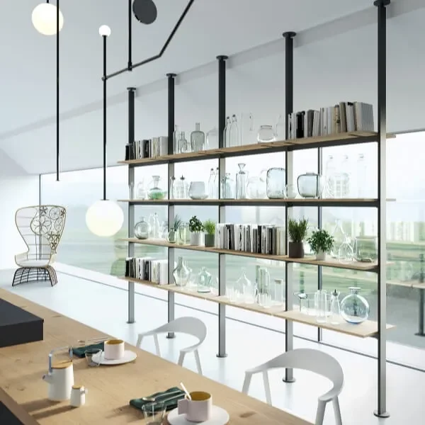 Modern dining area with a wooden table set with cups, utensils, and napkins. In the background, a large glass wall with a city view, and a tall open shelving unit with glassware, books, and potted plants.