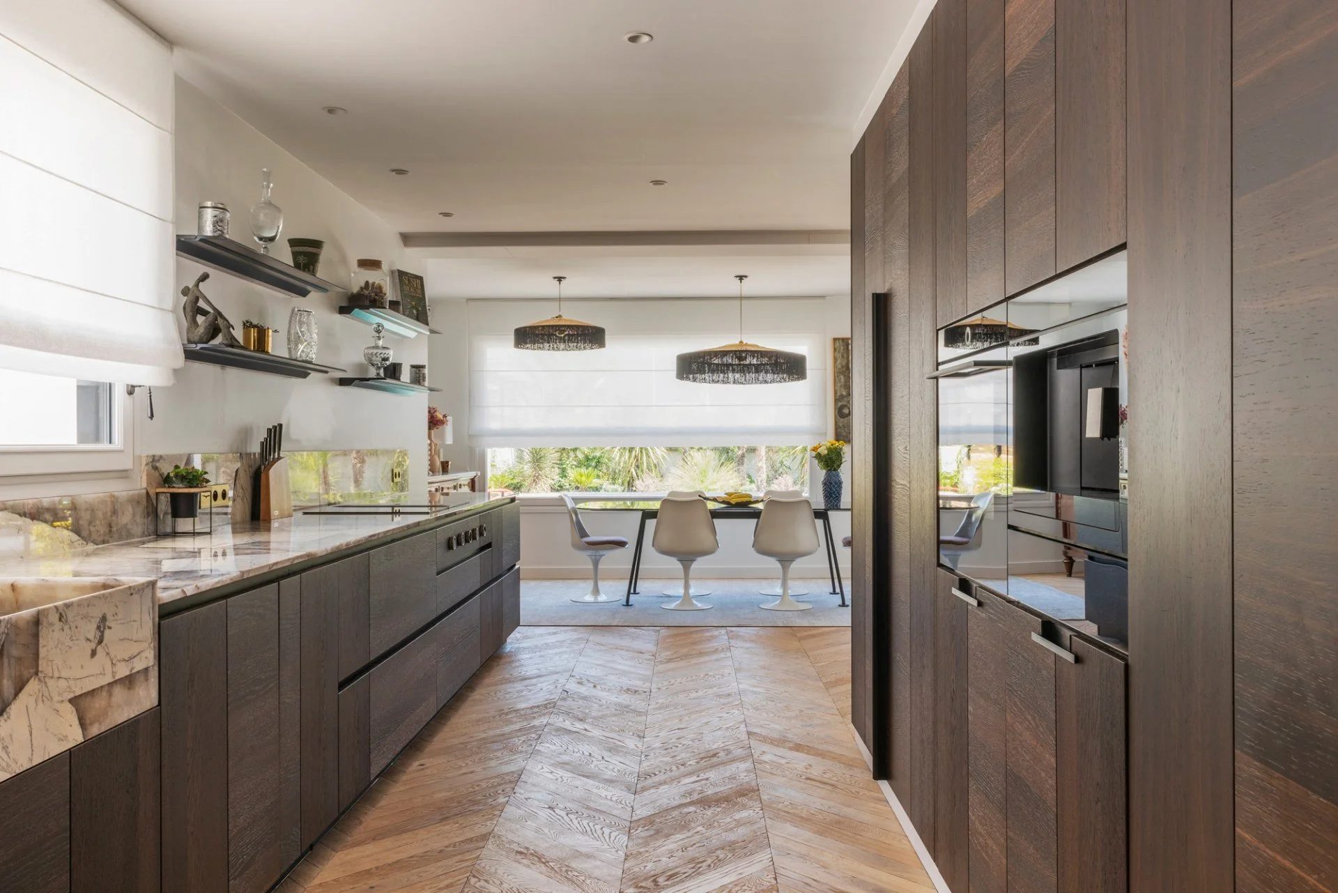 Modern kitchen with dark wood cabinets, marble countertops, and a dining area with white chairs and a table near large windows with blinds.