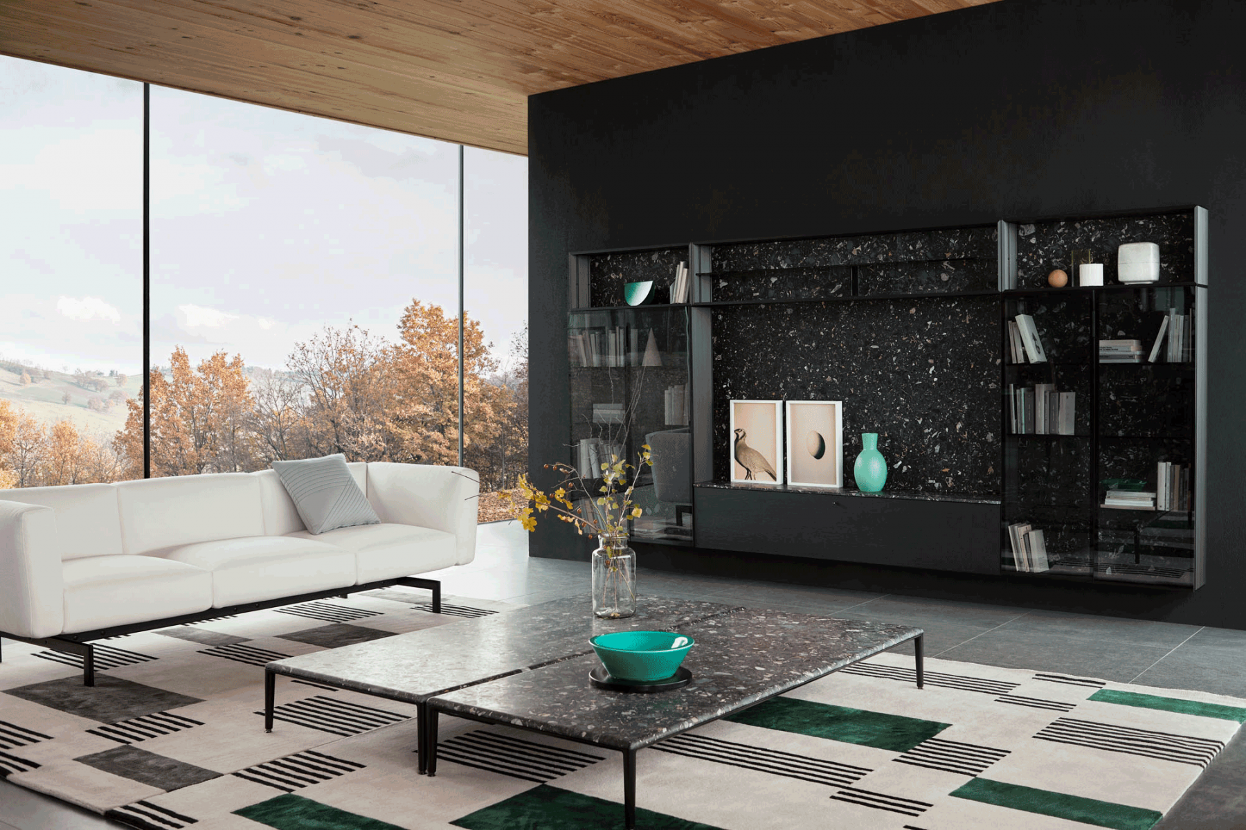 Modern living room with large floor-to-ceiling window showing trees with autumn foliage, a white sectional sofa with a gray pillow, a rectangular marble coffee table with a teal bowl, a black and white patterned rug, a black shelving unit with decora