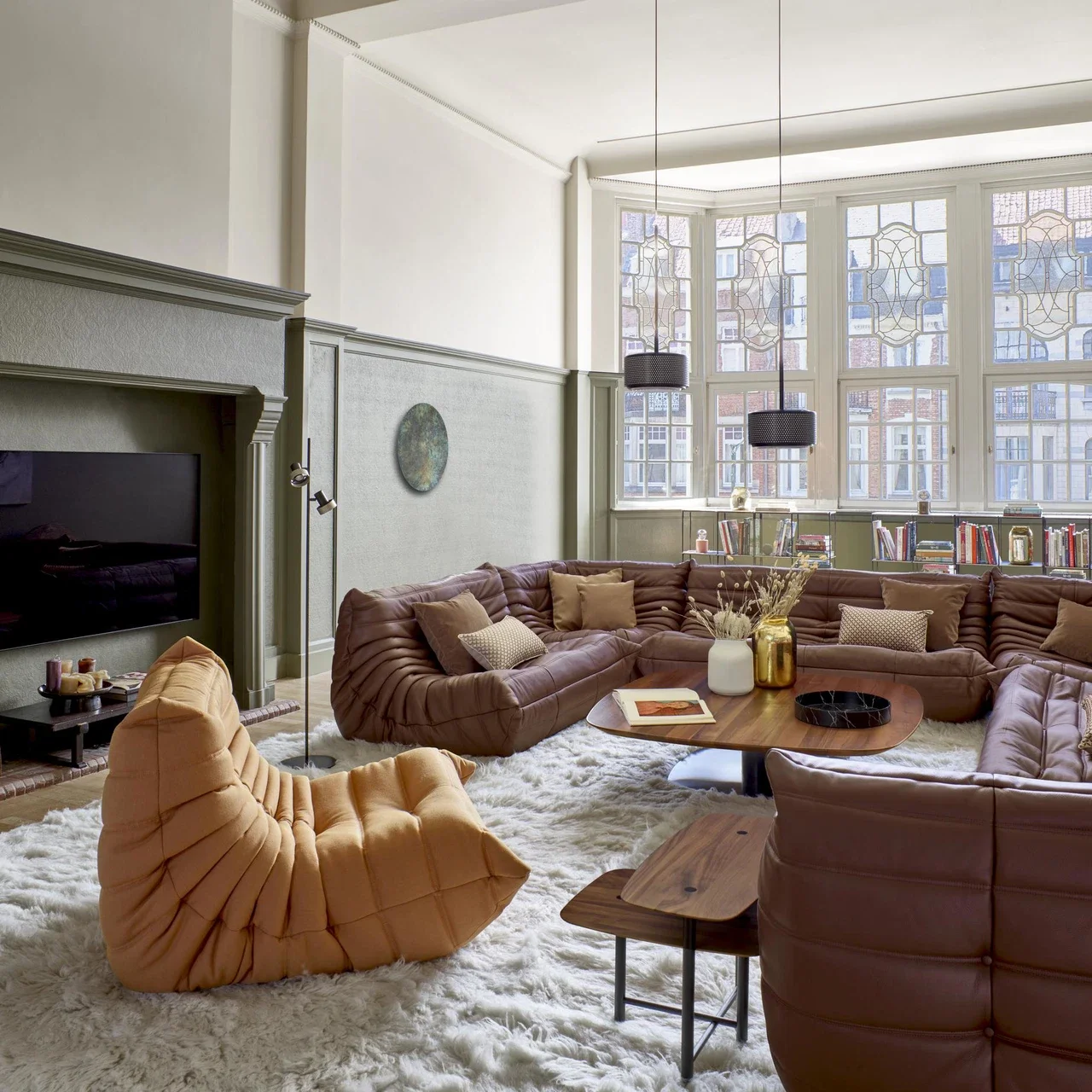 Living room with a large brown sectional sofa, tan armchair, white shag rug, wooden coffee tables, and large bay windows with stained glass. Decorative pillows, plants, books, and pendant lights are part of the decor.