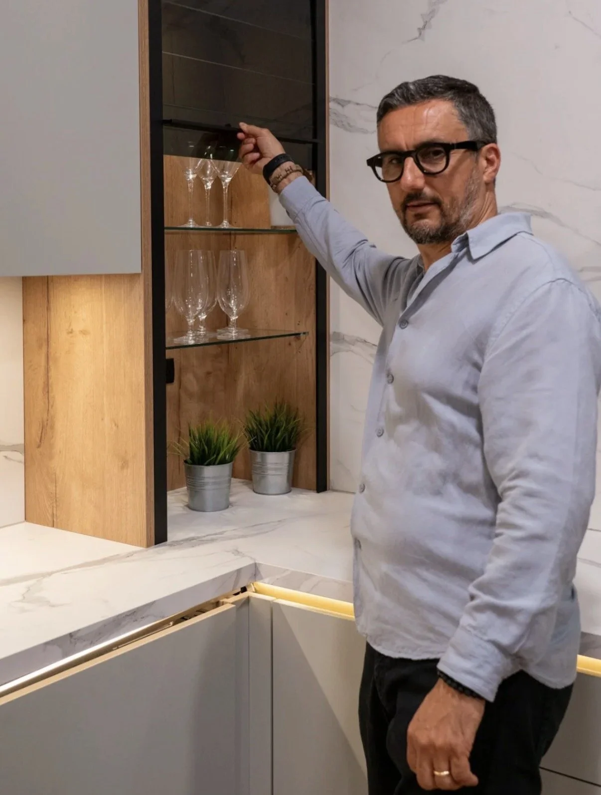 Man with black glasses and gray hair pointing at a display cabinet with wine glasses inside. The setting has a marble and wood interior with two potted plants on the counter.