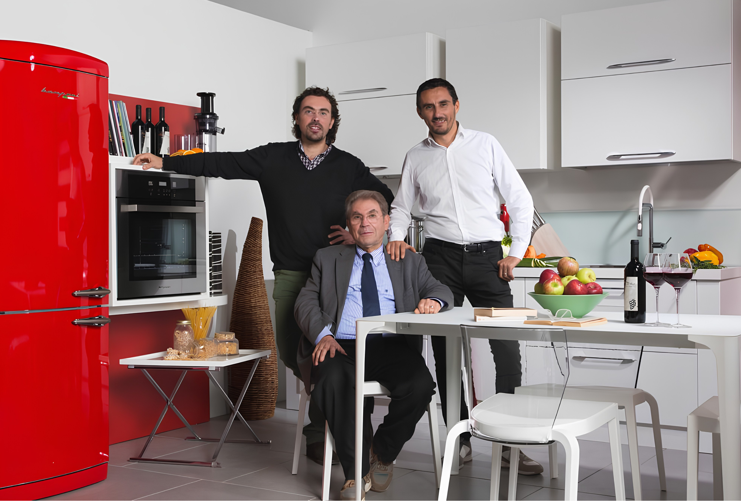 Three men in a modern kitchen with white cabinets and a red refrigerator. One man is sitting at a table with apples and wine, one is standing near a sink, and one with curly hair and a beard is leaning on a kitchen counter. There are various kitchen items and a cookbook on the counter.