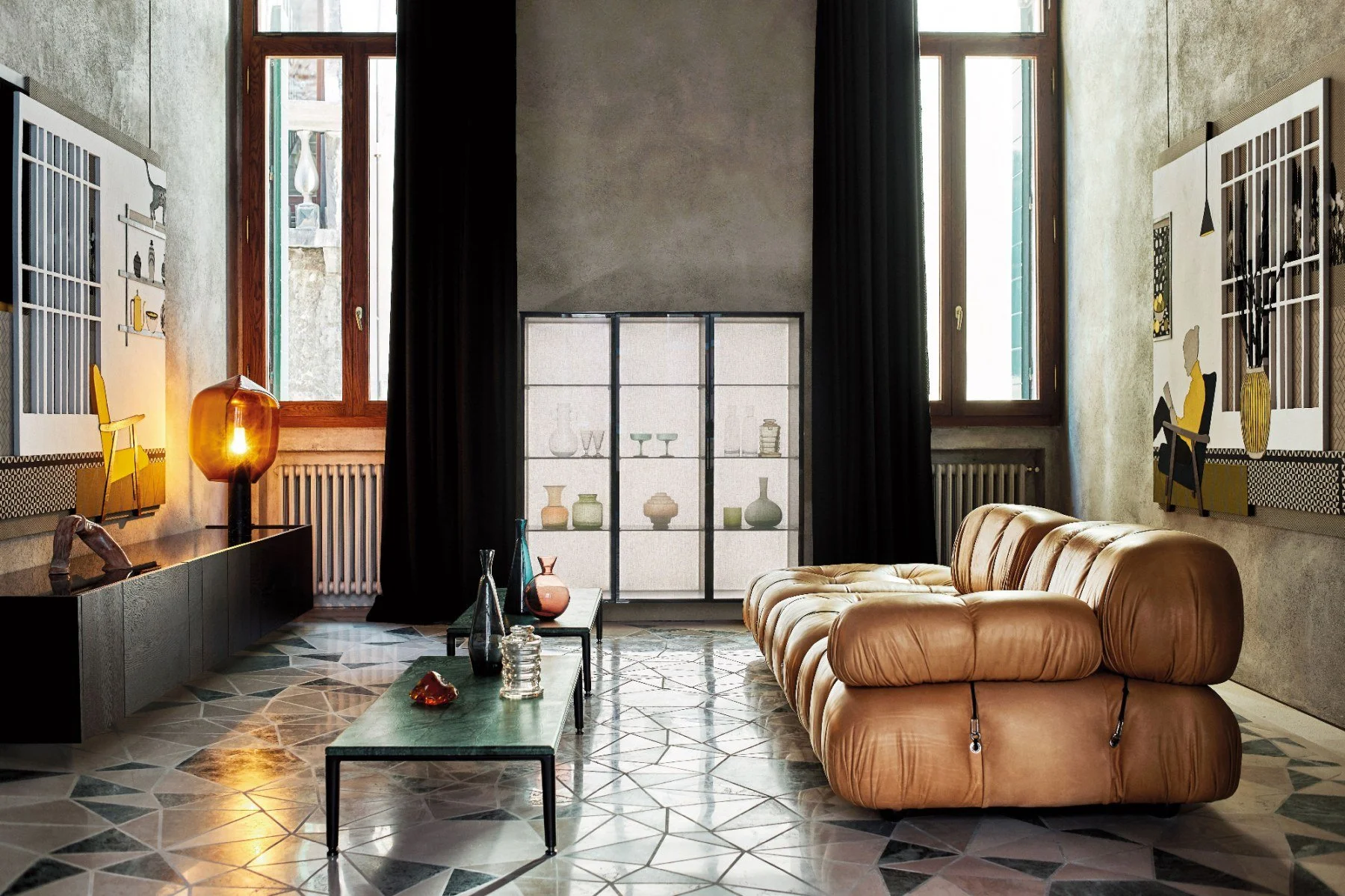 Living room with beige leather sofa, black coffee table with glass vases and decorative objects, shelving with vases, large windows with black curtains, patterned tile floor, wall art, and a warm lighting lamp.