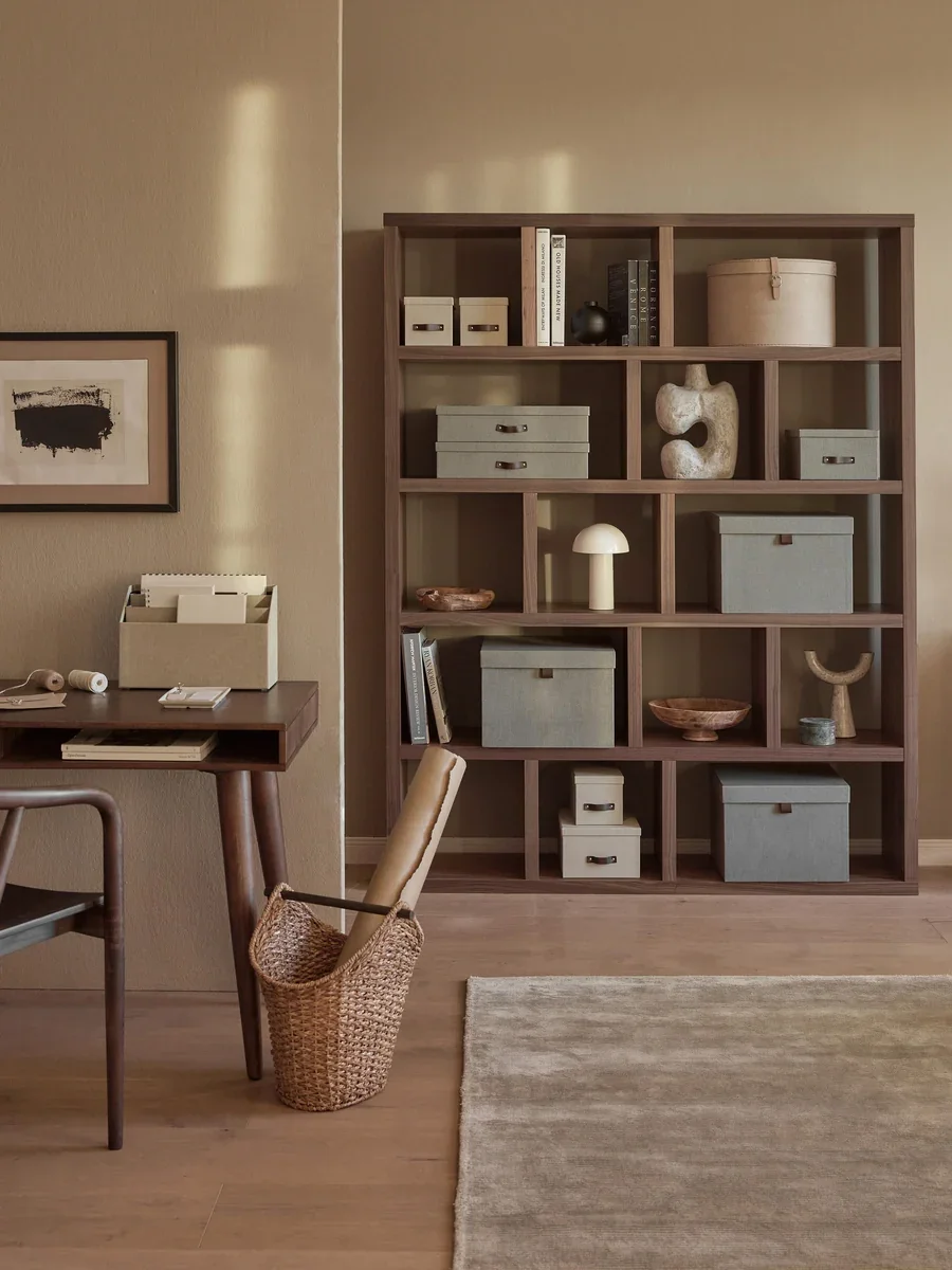 A cozy home office with a wooden bookshelf filled with decorative boxes, sculptures, and books, next to a wooden desk with notebooks and a wicker basket, beige walls, and a light wooden floor.