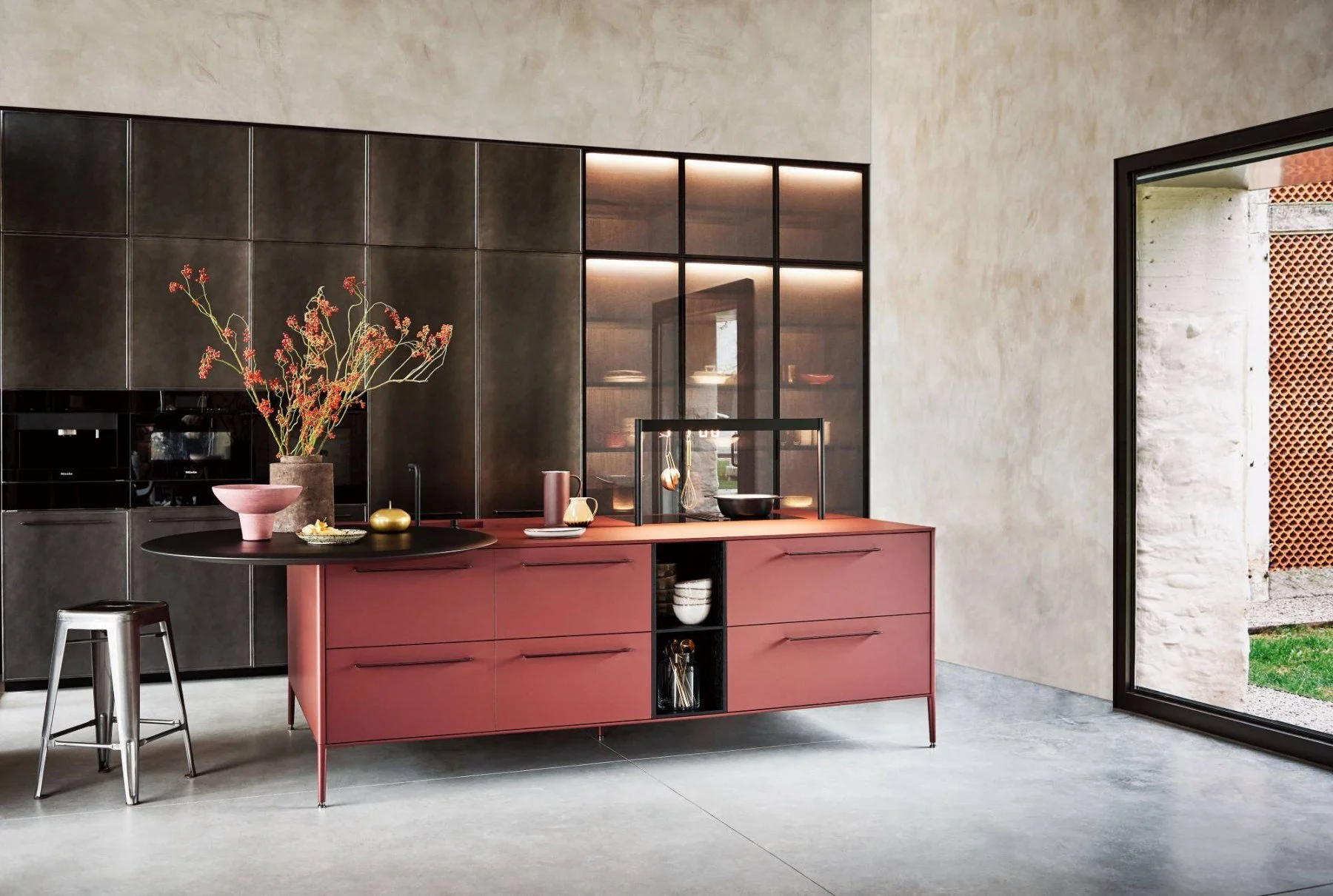 Modern kitchen with dark upper cabinets and a pink lower cabinet island, decorated with vases, bowls, and utensils; a black stool, a large window showing an outdoor brick wall and grass.