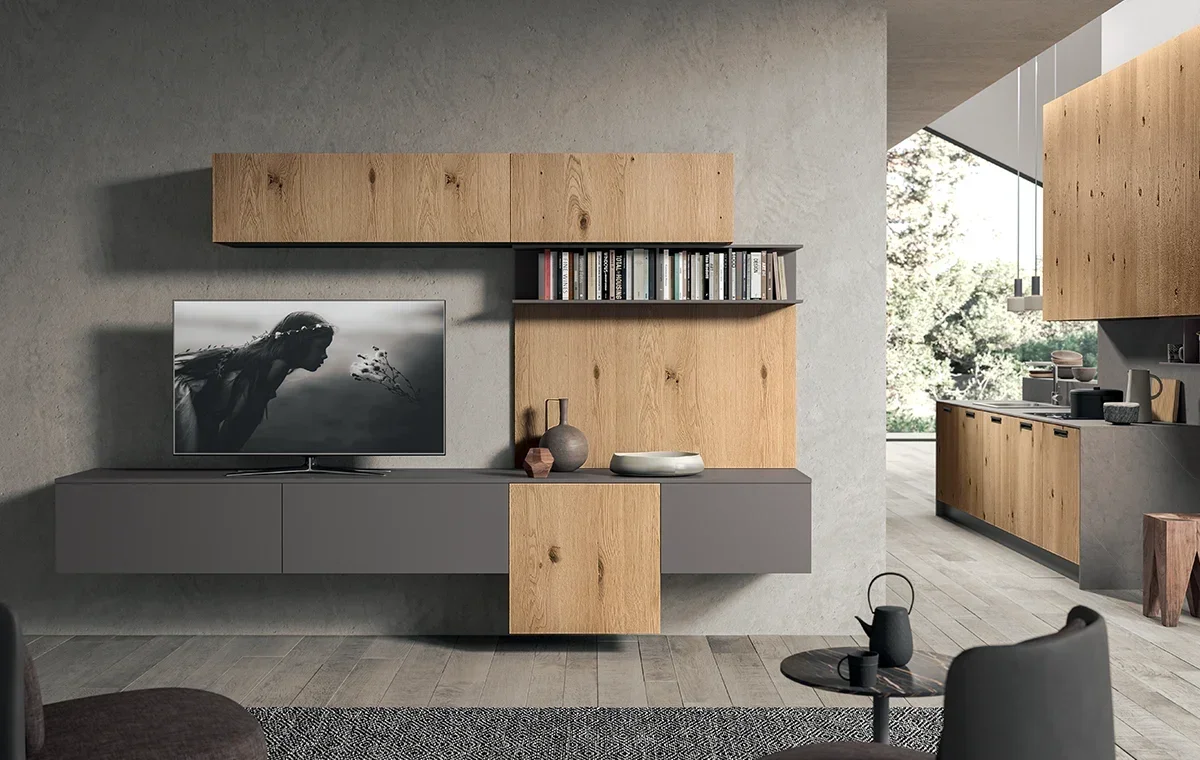 Modern living room with a wall-mounted TV displaying a black and white image of a woman, wooden and gray cabinetry, and a glimpse of the kitchen with wooden cabinets and a large window showing trees outside.