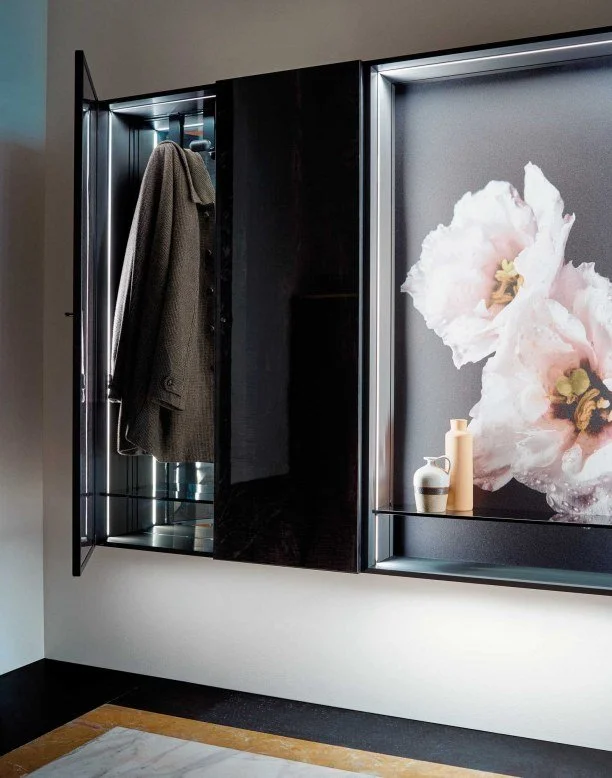 Modern black display cabinet featuring a coat hanging inside and decorative vases on illuminated shelf with flower artwork in the background.