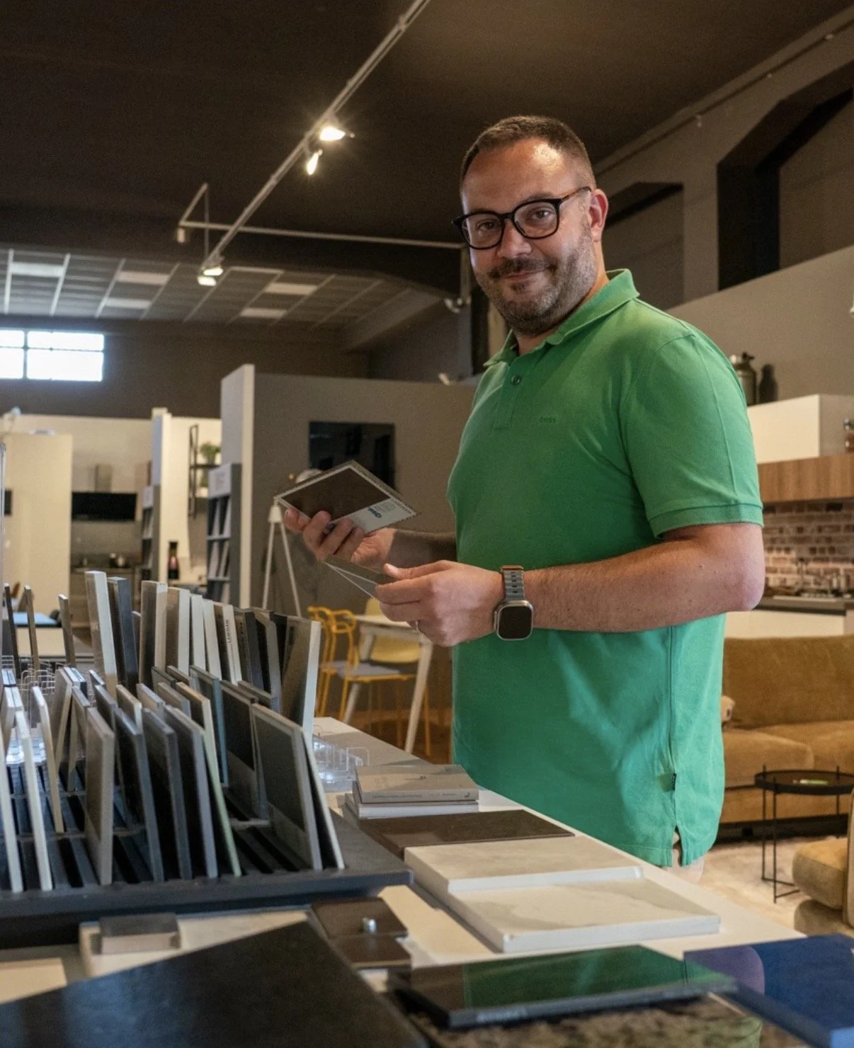 A man with glasses and a beard wearing a green polo shirt and smartwatch is browsing through samples or swatches at a store that sells home decor or furniture.