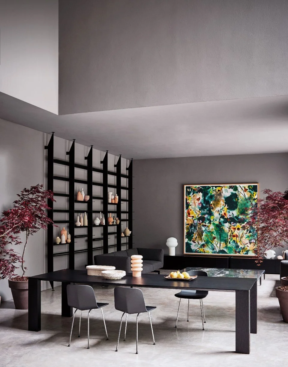 Modern living room with a black dining table, gray chairs, potted red-leafed trees, abstract colorful wall art, and decorative vases.