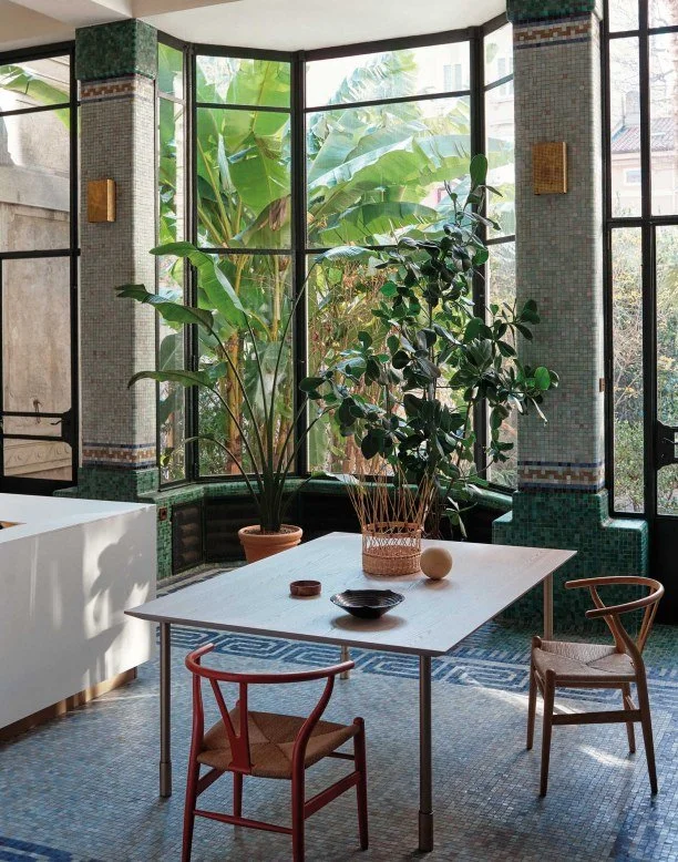 Bright sunlit room with large windows overlooking a lush garden. Inside, a white table with decorative bowls and vases, two wooden chairs, and tropical plants in pots.