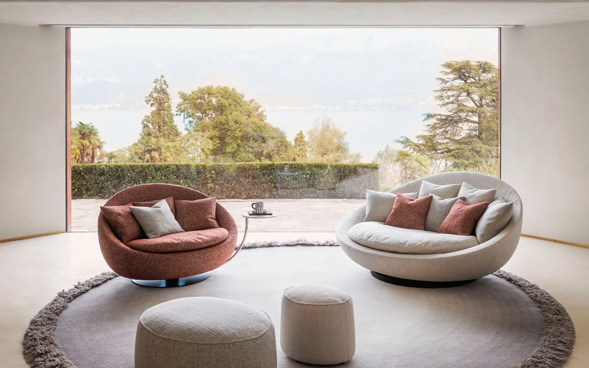 Modern living room with two curved armchairs, one red and one light gray, with cushions, in front of a large window overlooking a lush green landscape with trees, water, and mountains in the background.