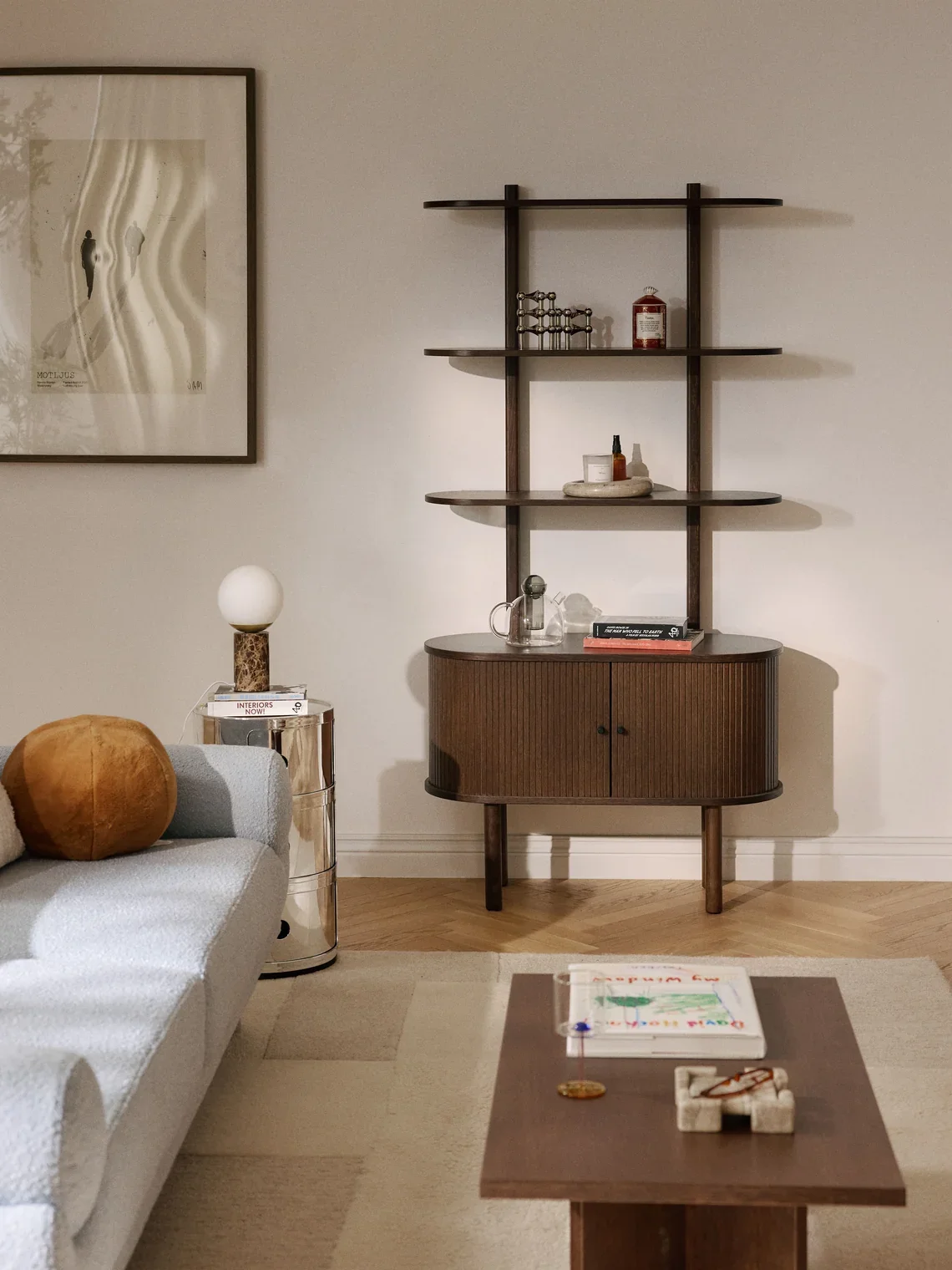 Living room with a light-colored sofa, a round side table with a lamp, and a wooden shelving unit with decorative items and books against a white wall. There is a framed picture on the wall.