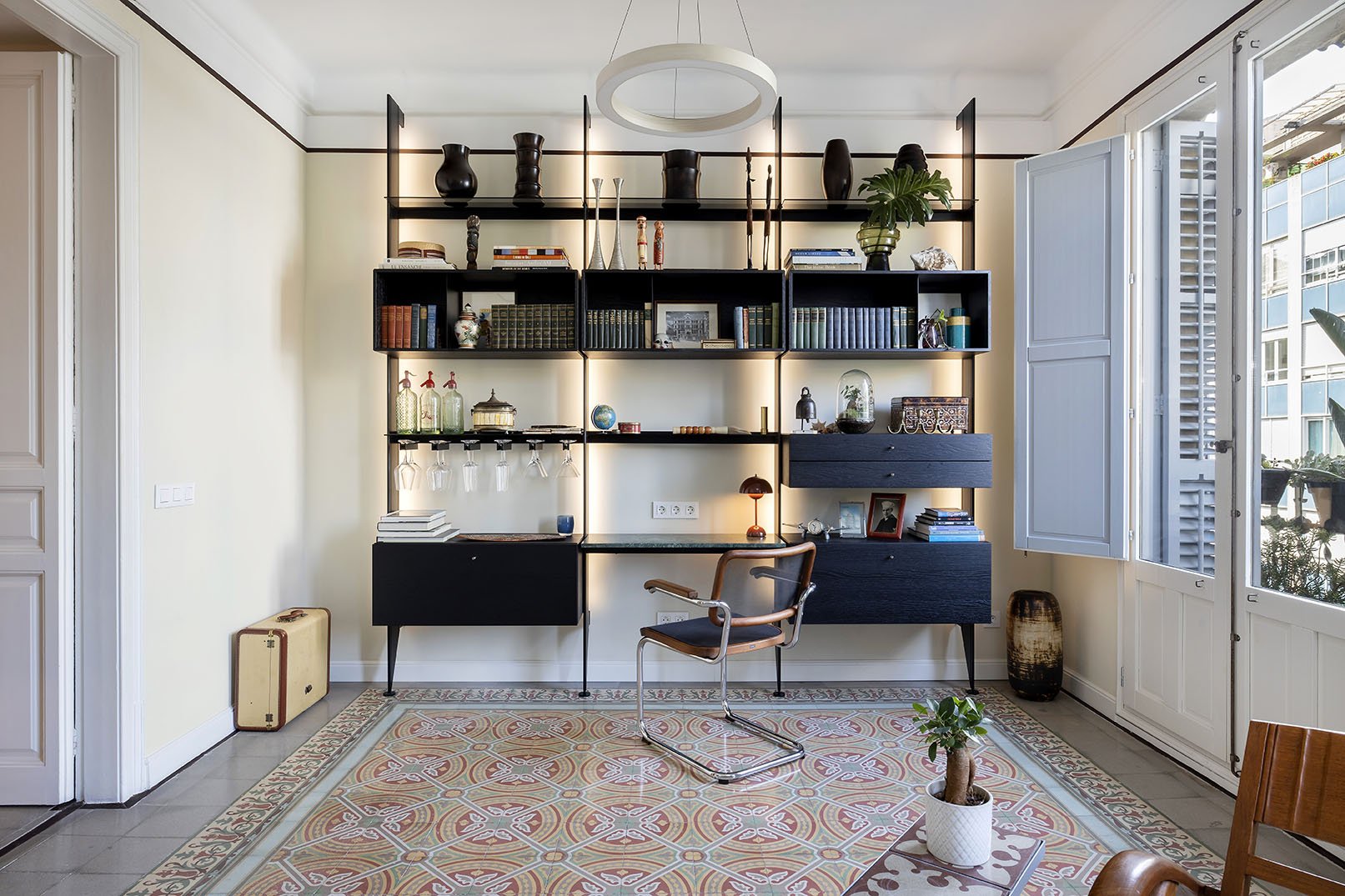 Interior view of a cozy home office with a black and wooden bookshelf wall unit, a modern chair, a patterned rug, a desk with a small lamp, and large windows letting in natural light.