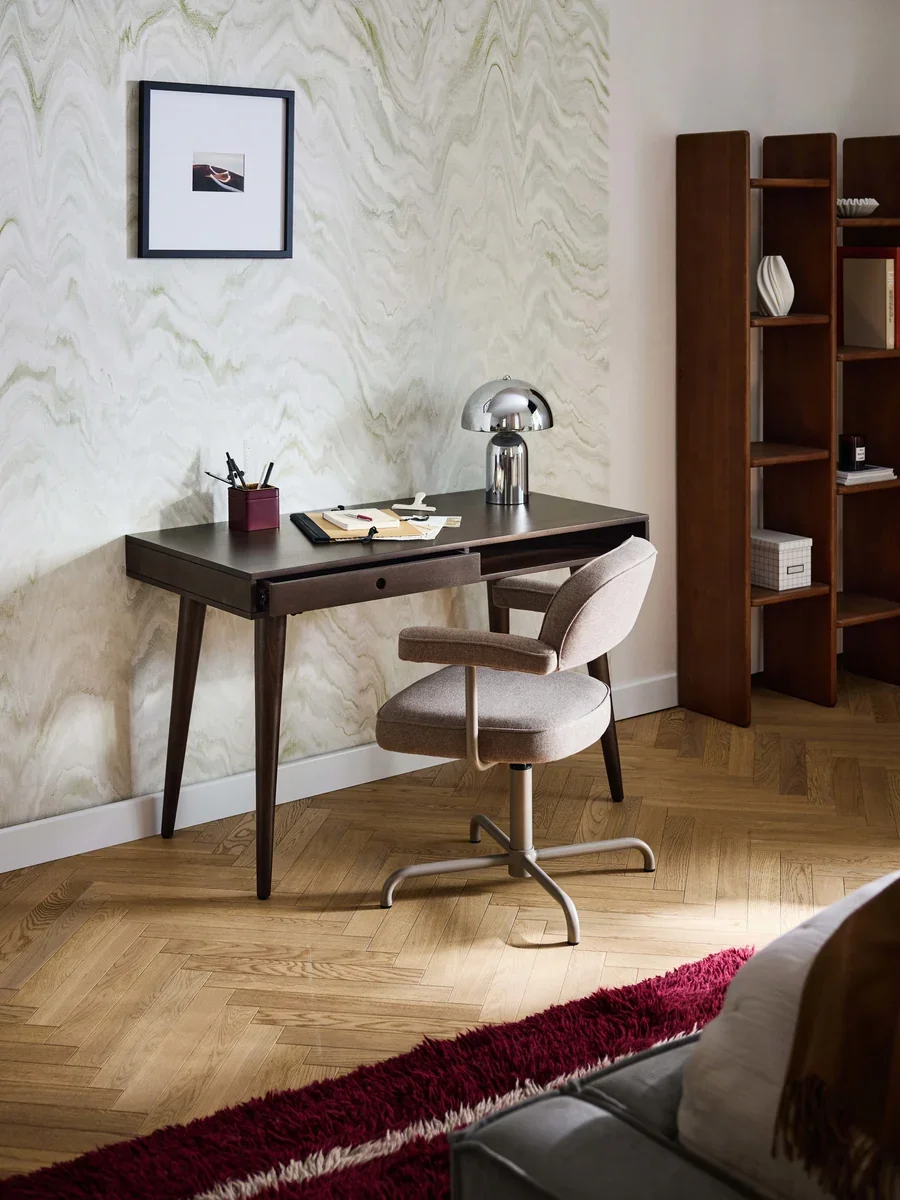 A cozy home office with a dark wooden desk, beige swivel chair, silver desk lamp, framed photograph on the wall, wooden bookshelf, and a red and white rug on a parquet floor.