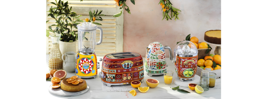A colorful breakfast spread with juice, oranges, and cakes, featuring vibrant, patterned kitchen appliances and decor with lemon slices and greenery.