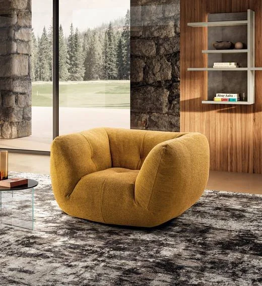 Modern living room with a mustard yellow armchair, a round glass coffee table, a textured gray area rug, large floor-to-ceiling windows showing a forest, and a wall with stone and wood paneling featuring a white wall shelf with books and decorative i