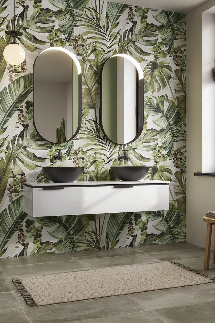A modern bathroom with tropical leaf-patterned wallpaper, two black vessel sinks on a white floating vanity, and two vertically oval mirrors above each sink.
