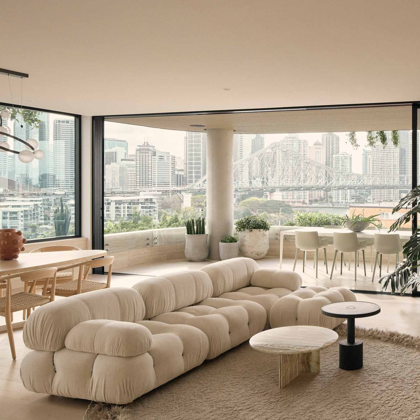 Modern living room with large white sectional sofa, round marble side table, and mountain view through floor-to-ceiling windows. Flowers and plants decorate the space.
