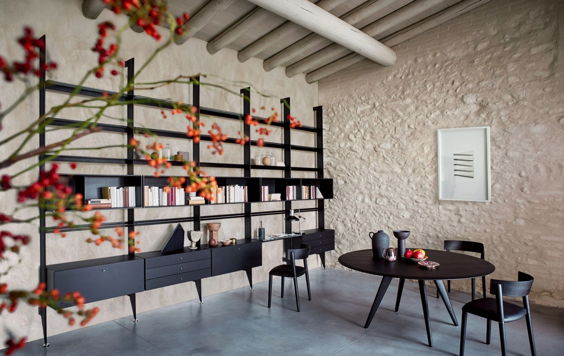A modern dining room with a round black table set with vases and fruit, surrounded by black chairs. There is a black wall shelving unit with books and decorative items, and textured light-colored stone walls. A white framed art piece hangs on the wal
