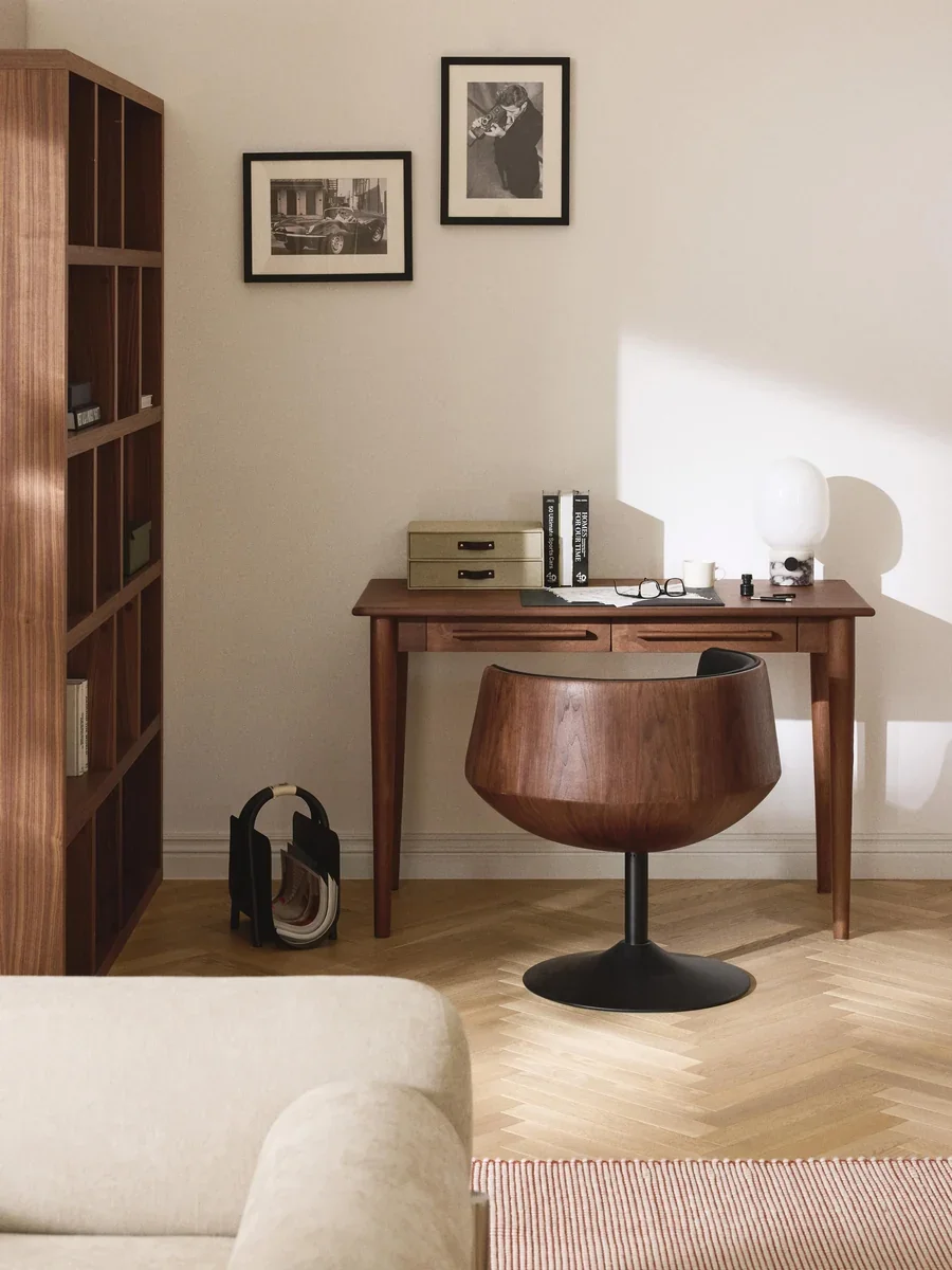 Home office with a wooden desk, a brown swivel chair, framed black-and-white photos on the wall, a white table lamp, books, glasses, and decorative items.