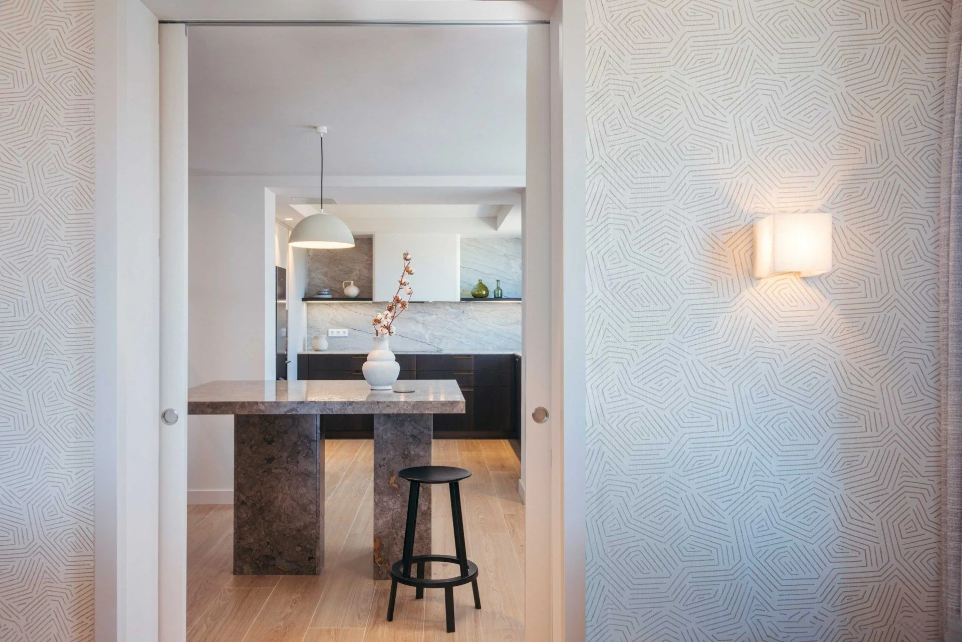 View through partially open white sliding door to a contemporary kitchen with a small black stool, marble counter, decorative vases, and patterned wallpaper on the wall.