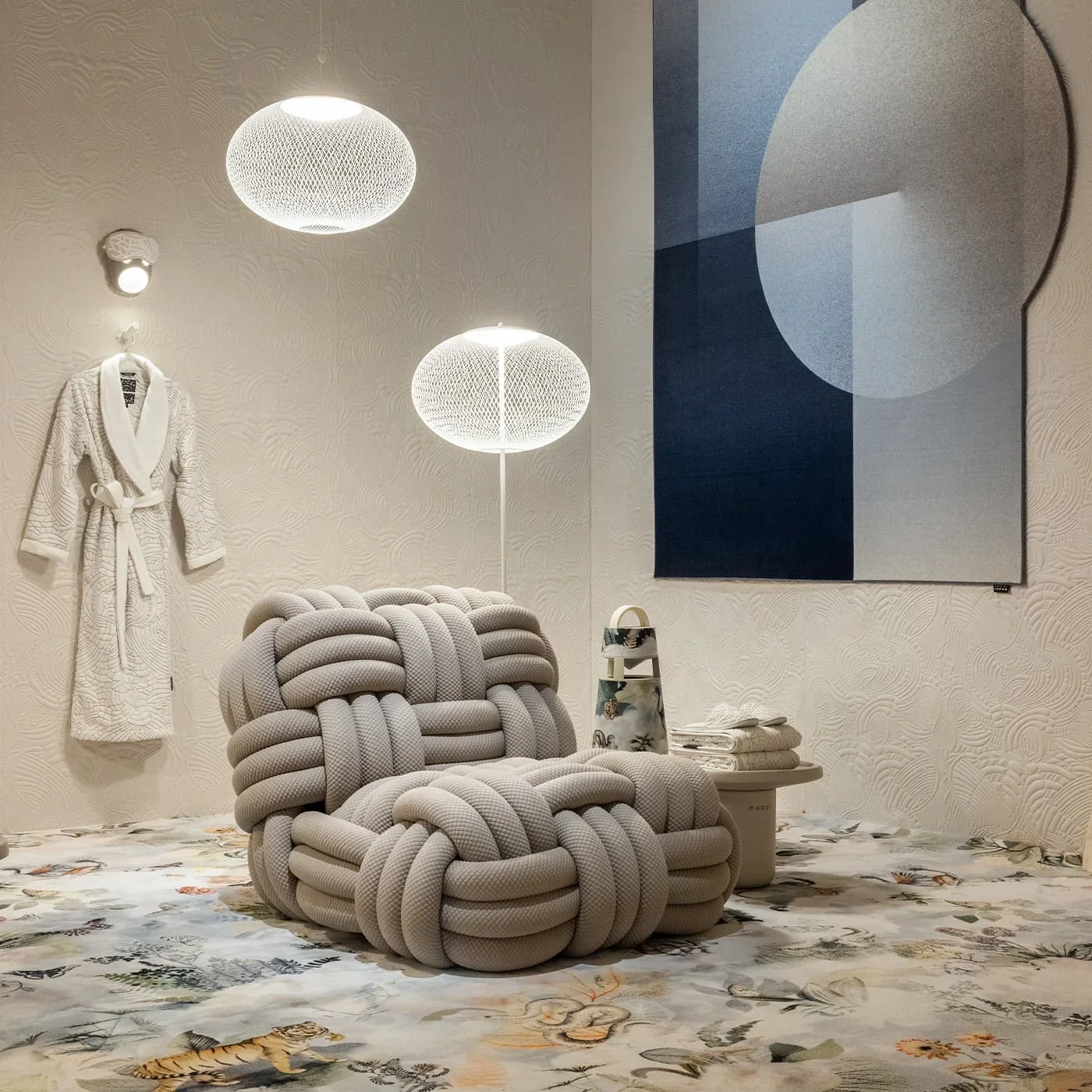 Modern interior with a textured white wall, a gray knotted sofa, a white table with folded towels, a colorful ceramic pitcher, an abstract blue, gray, and beige wall art, and patterned floor and wall lighting.