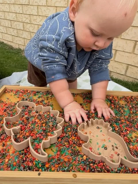 Young child playing with colored beans and cookie cutter shapes on a sandbox outdoors.