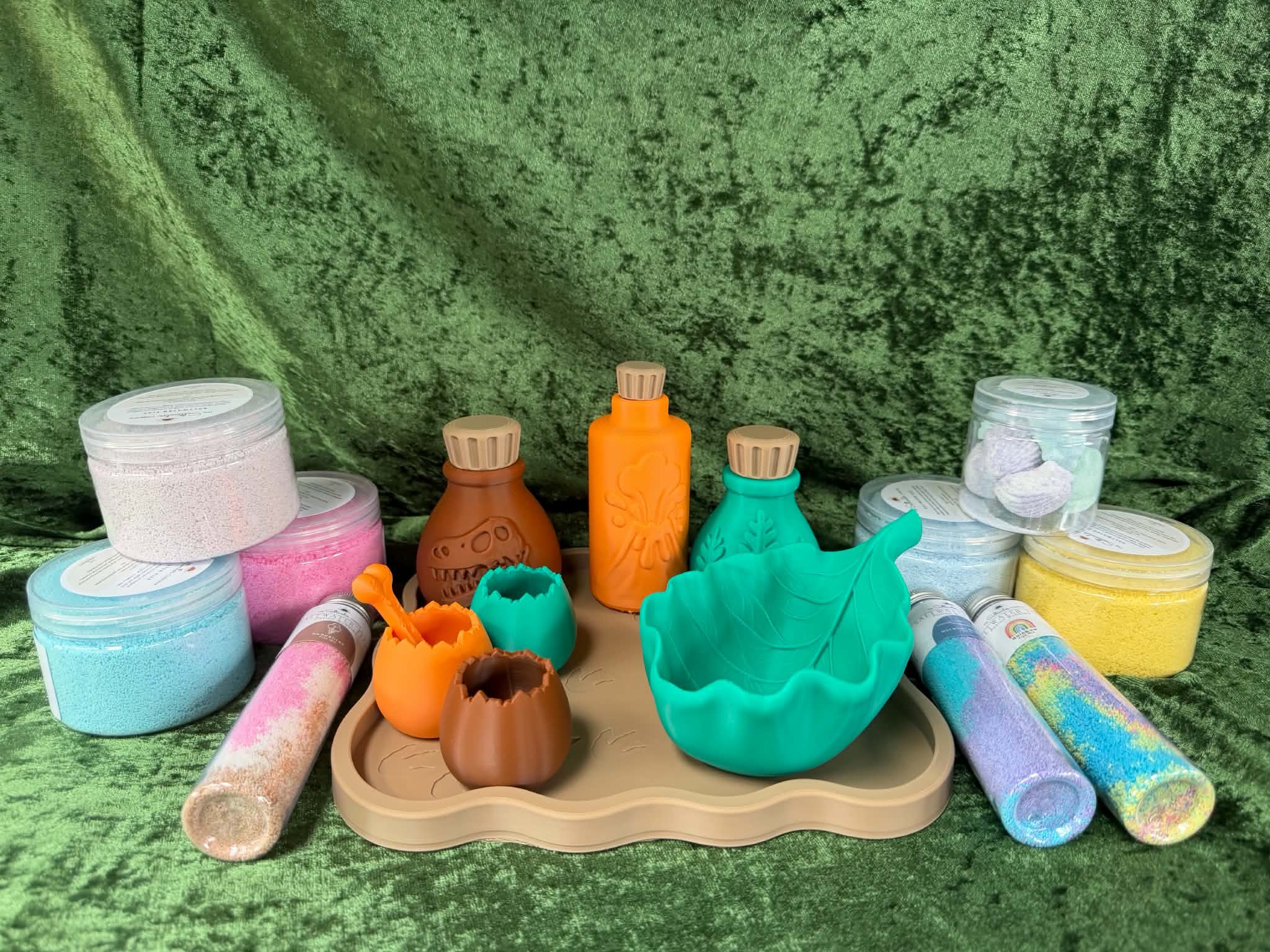 Colorful sensory and craft materials displayed on a green fabric background, including jars of textured materials, small bottles, silicone molds, and rolled and cut foam sheets.