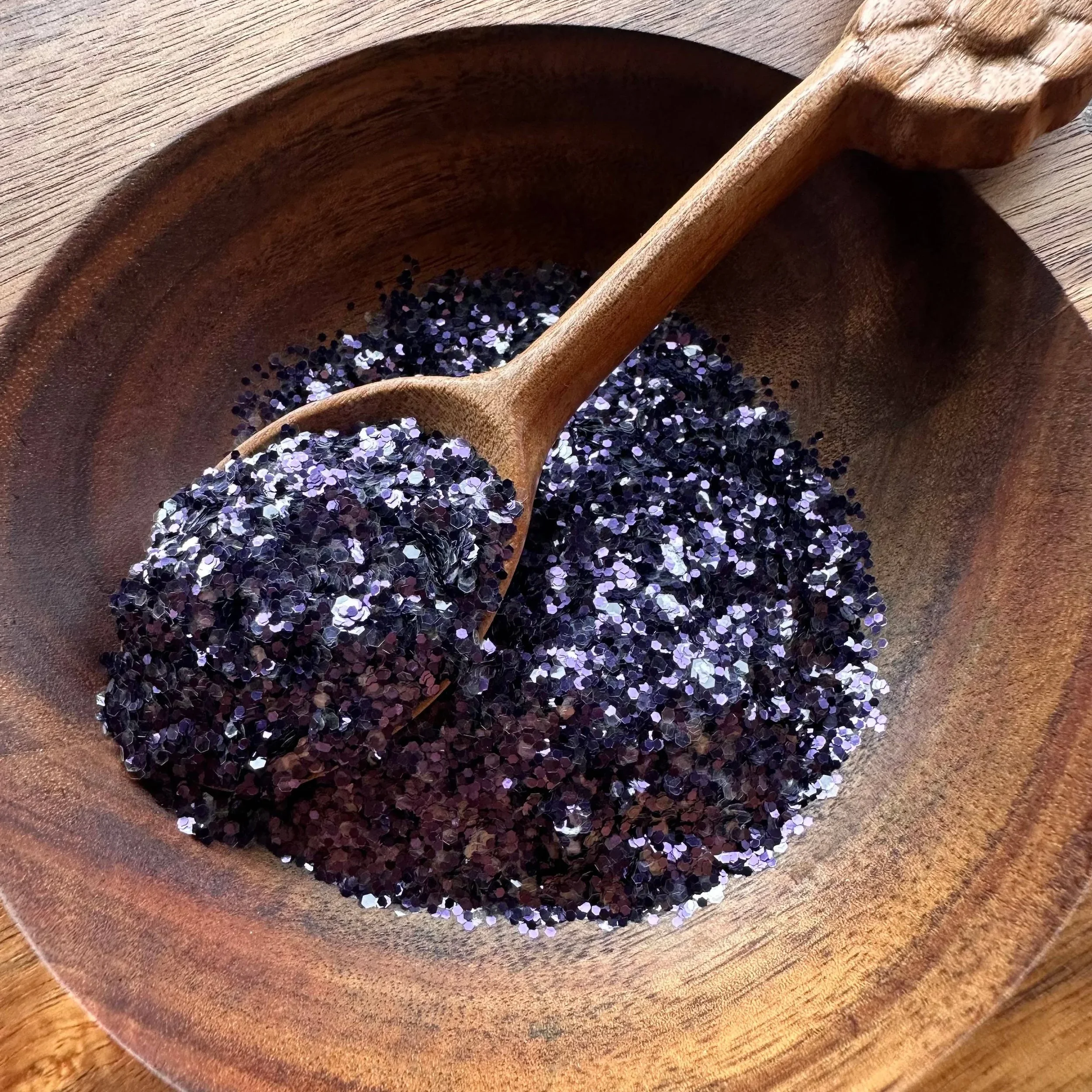 A wooden bowl filled with purple and black glitter, with a wooden spoon resting inside, on a wooden surface.