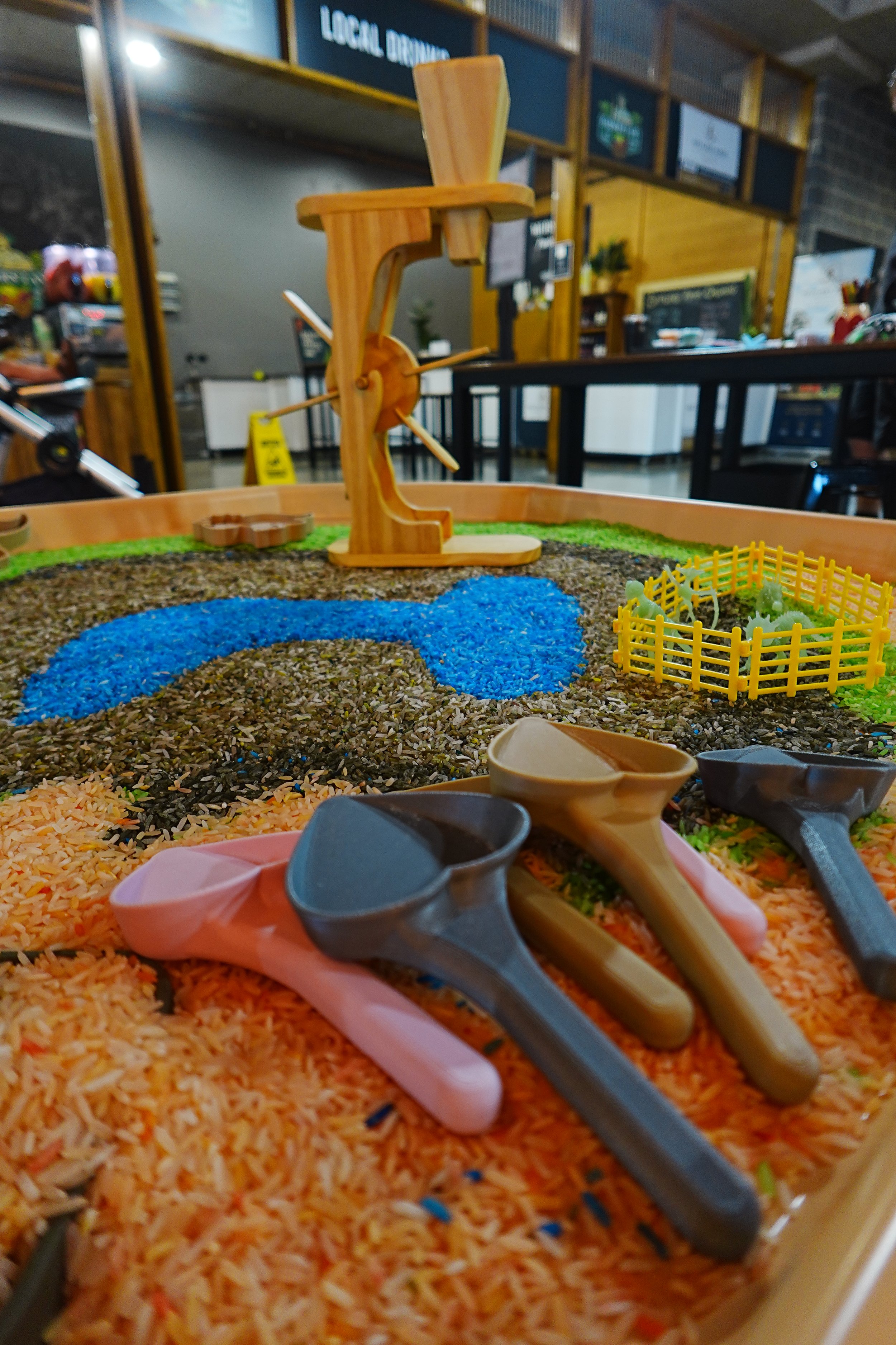 A display of colored rice and small plastic scoops on a table, with a small wooden chair model in the background, inside a restaurant or cafe setting.