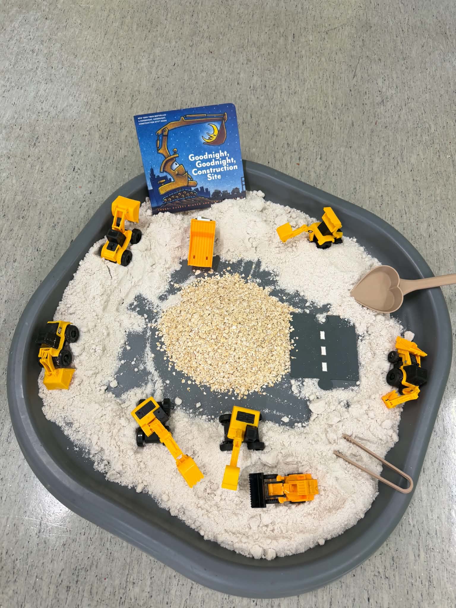 Play area with toy construction vehicles, sand, and a book titled 'Goodnight, Goodnight, Construction Site' on a gray plastic surface.