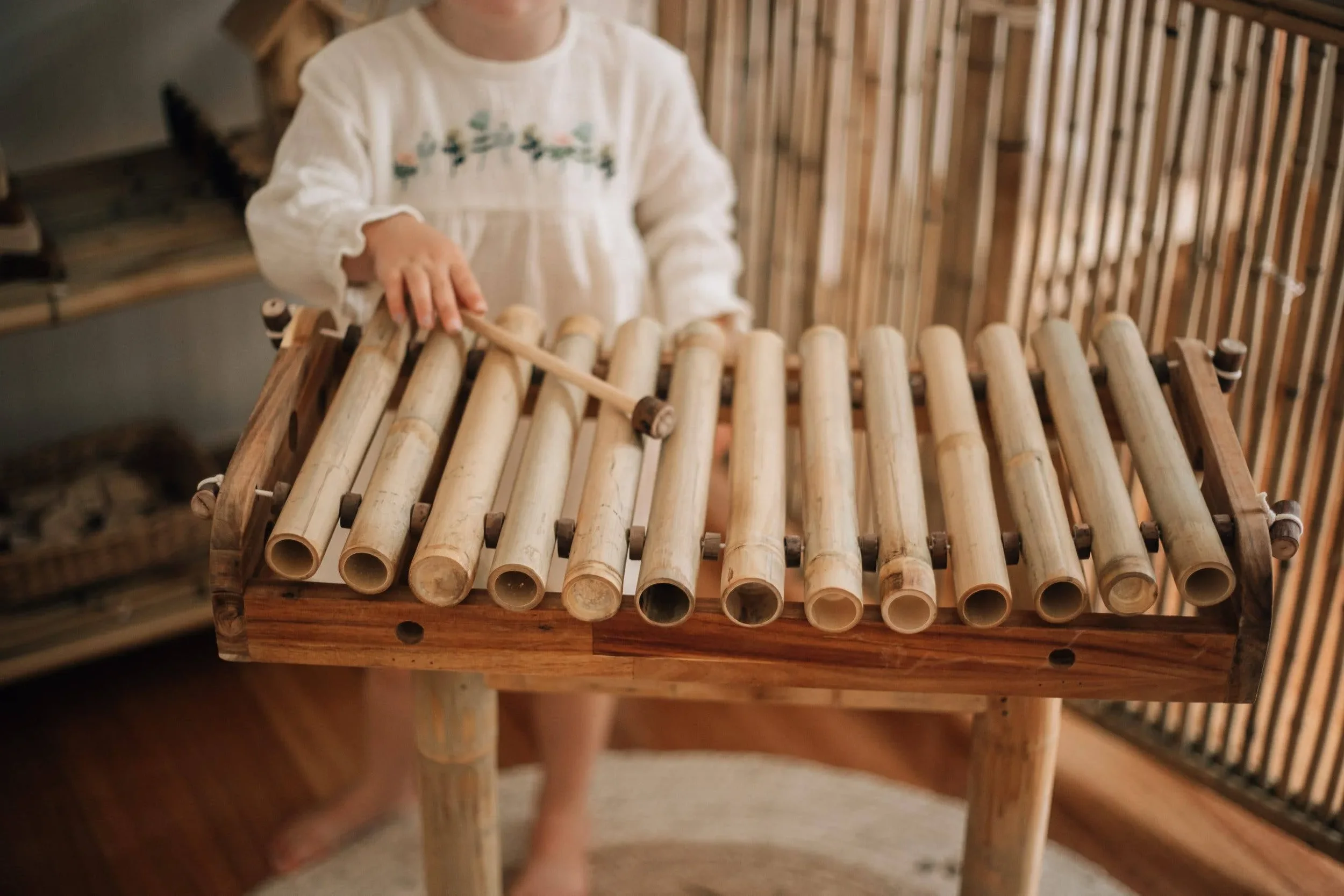 Child playing a wooden xylophone made of bamboo tubes, holding a mallet, in a room with wooden furniture.