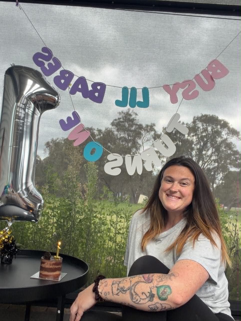 A woman with long hair and tattoos sitting at a table with a small cake and a candle, smiling, during a birthday celebration. There are birthday balloons and a banner behind her.