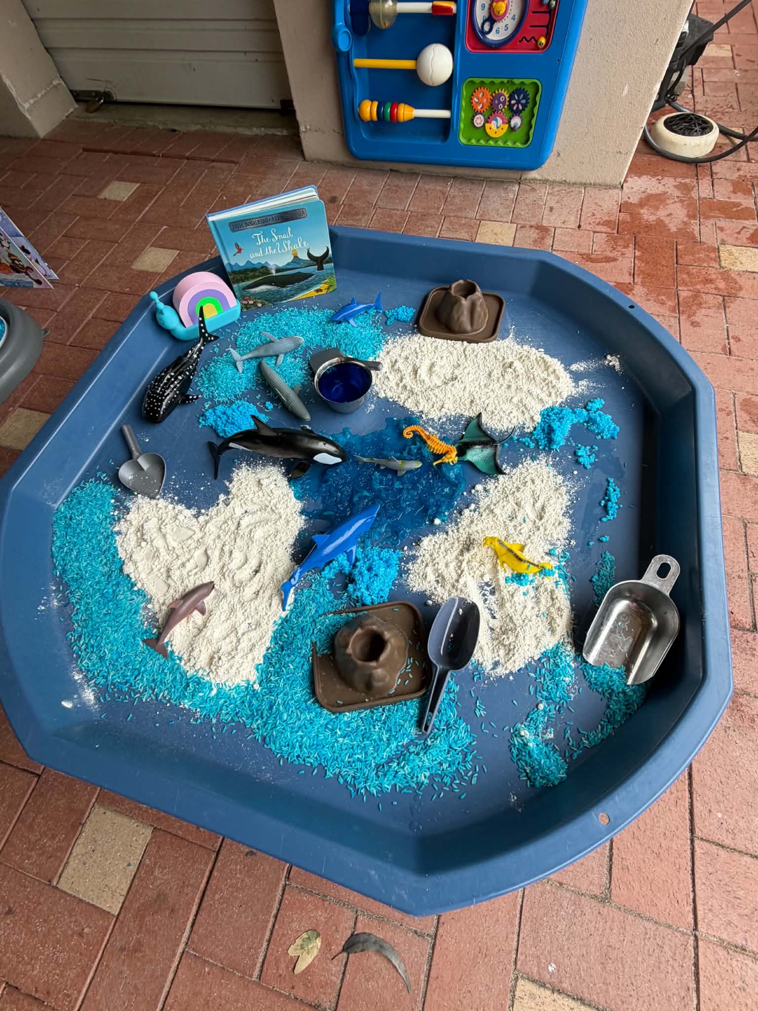 Blue sandbox with plastic toys, small animal figurines, a book titled "The Small and the Whale," and shredded blue and white material, situated on red brick patio.