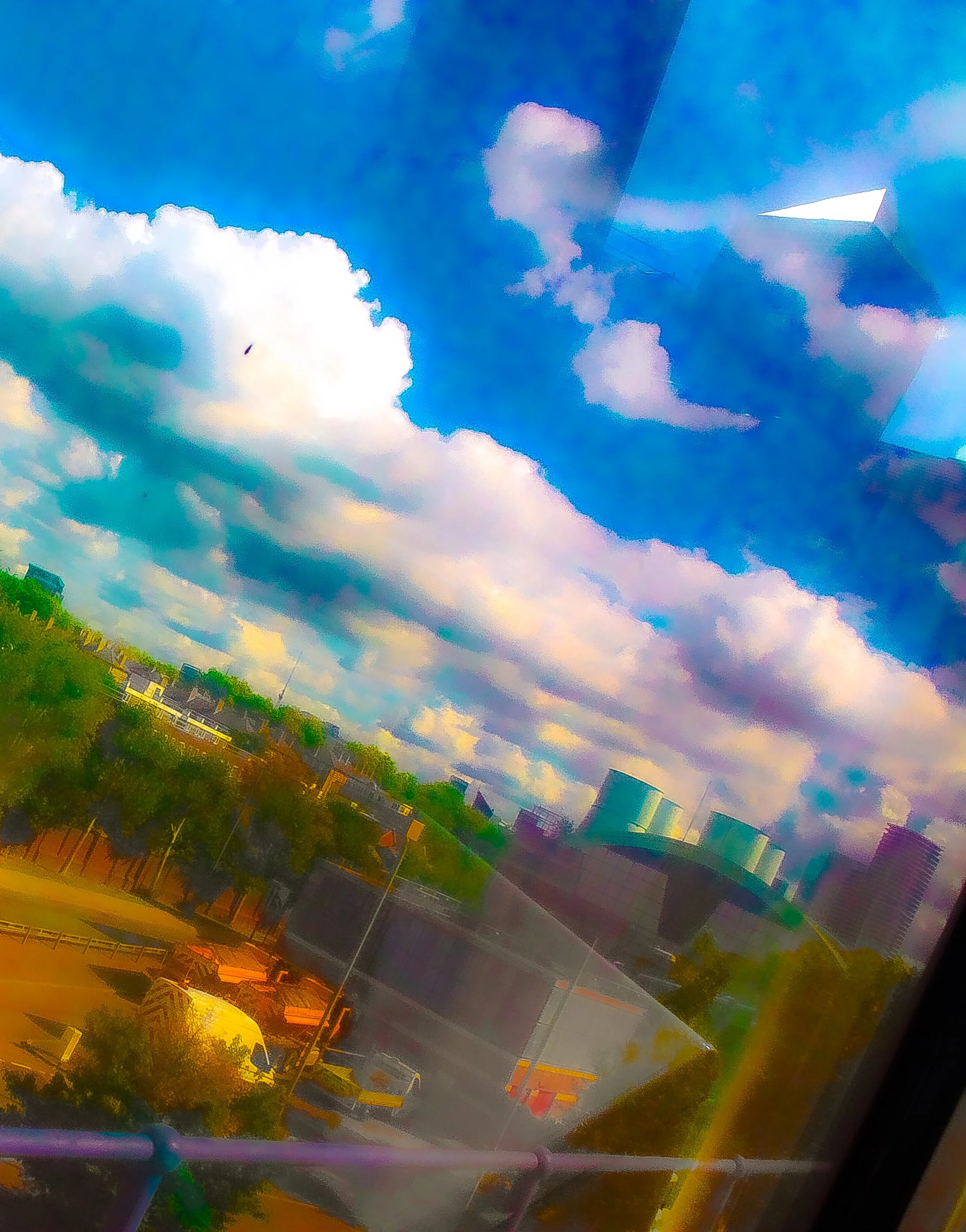 A colorful abstract photograph with a bright blue sky, clouds, and reflections on a glass surface, with hints of a neighborhood and trees below.