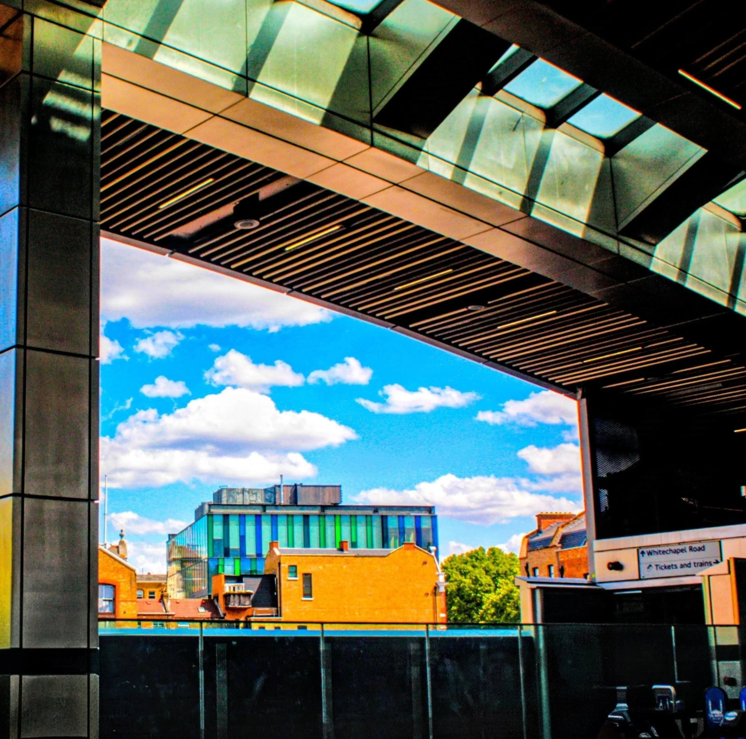 View through a glass window showing a blue sky with clouds, urban buildings with colorful rooftops, and a street scene with cars and a sign for Whitechapel Road.