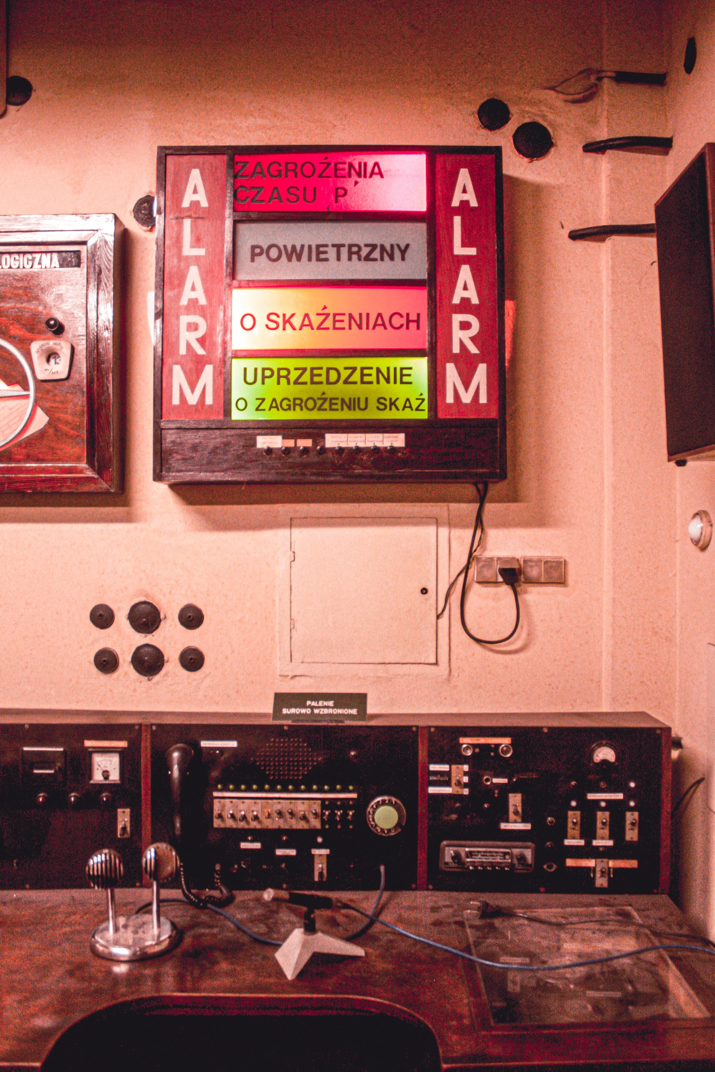 An old control room with various electronic devices, a vintage microphone on a stand, and a wooden desk in the foreground. A warning sign on the wall states 'FALENIE SUROWO WZABRONIONE'