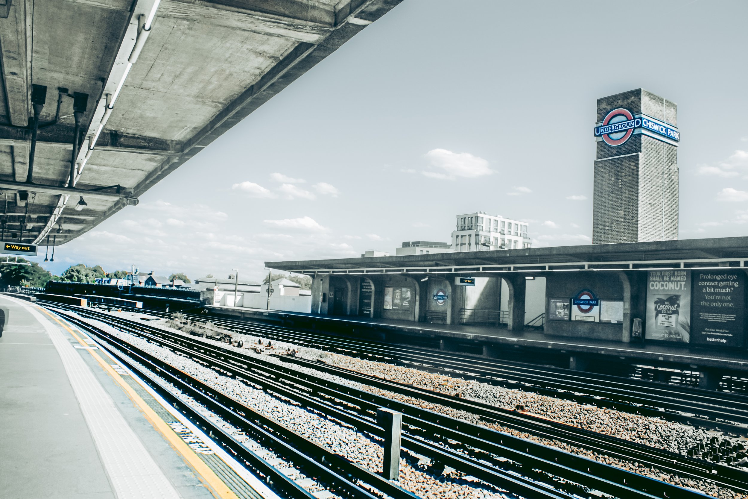 View of Chiswick Park underground station platform with train tracks, an overpass with pipes underneath, and a cityscape in the background under a partly cloudy sky.