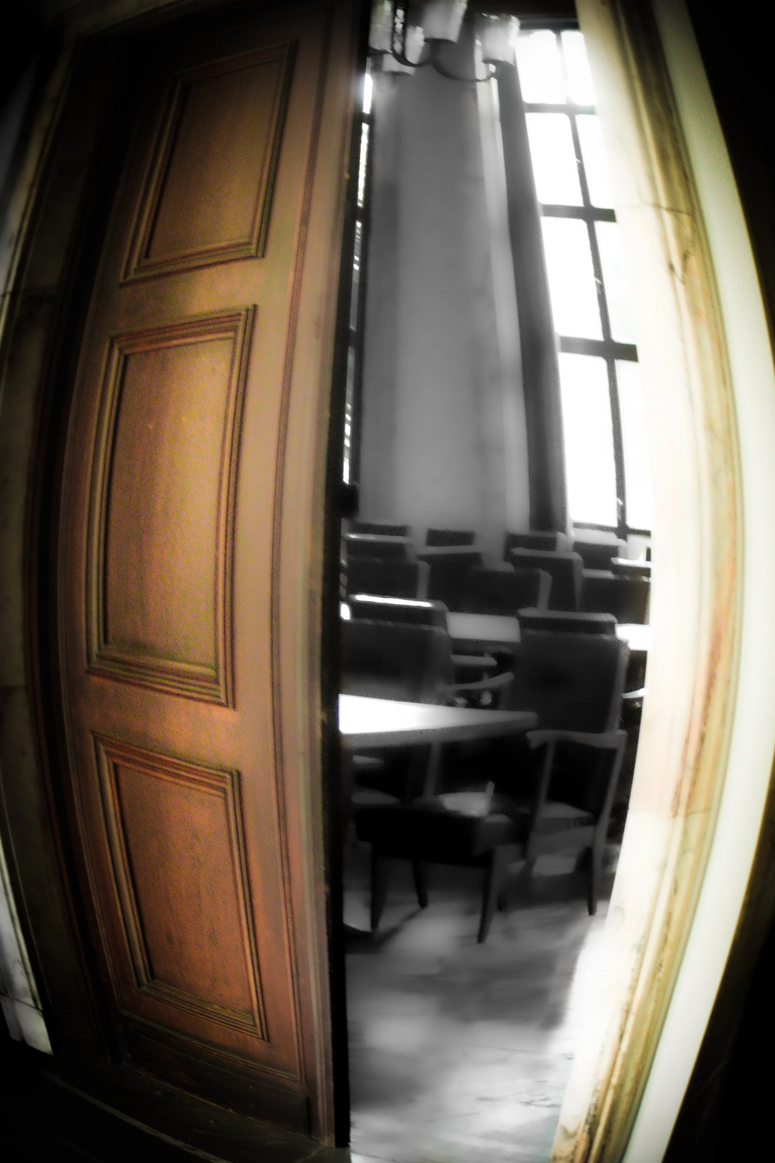 View through a slightly open wooden door with a carved panel design into a room with multiple chairs arranged around a windows, allowing sunlight to illuminate the space.