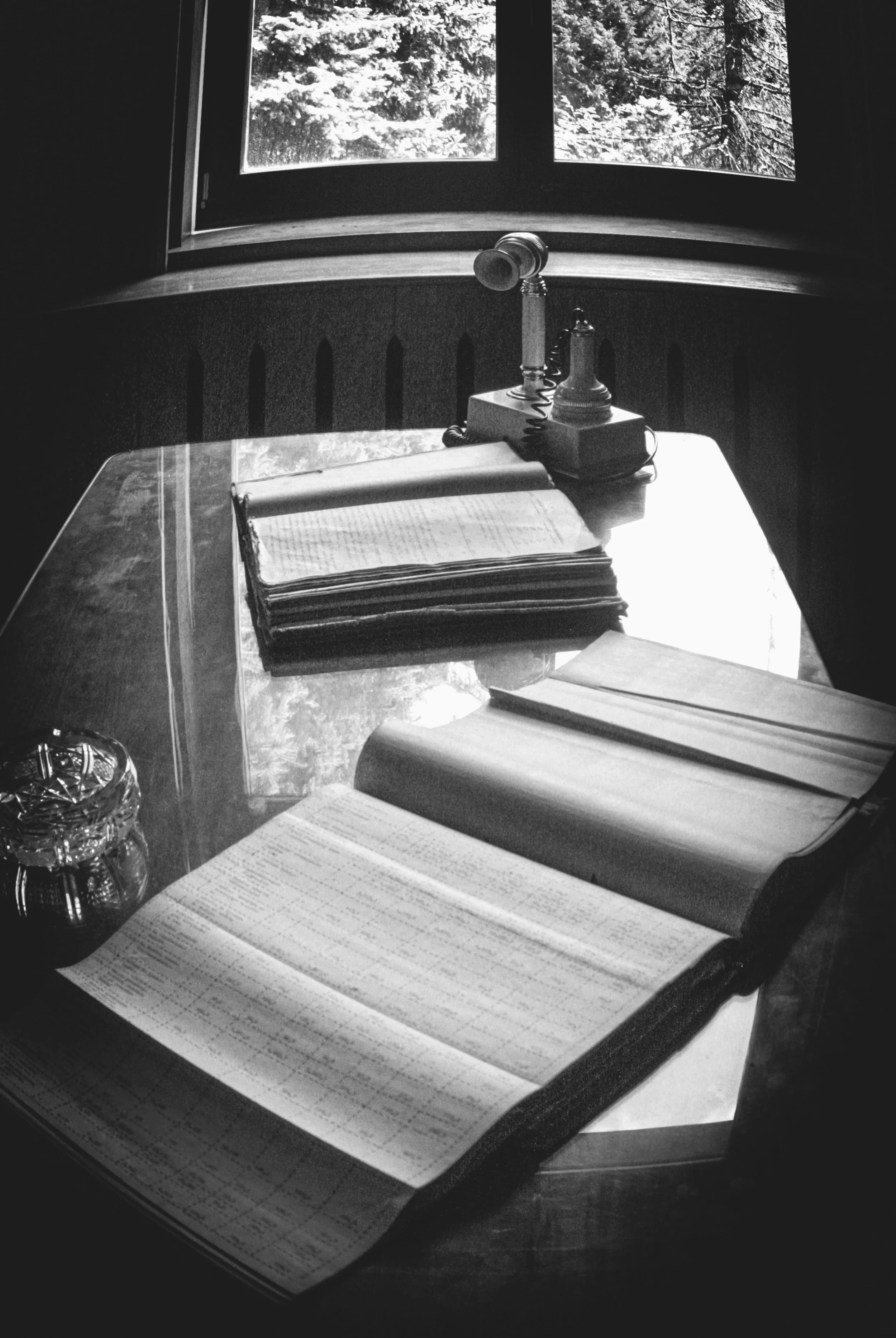 Black and white photo of a wooden desk with open books, notebooks, and a glass container with a lid. A vintage rotary phone is placed on the desk near the window, which has a view of snow-covered trees outside.
